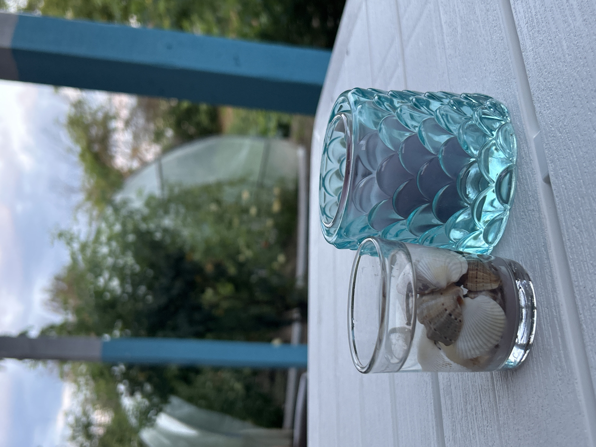 On a white, slatted table, two candle holders are displayed. To the left, a clear glass votive holder contains sand and several seashells. To the right, a turquoise, scaled glass candle holder holds a dark-colored candle. The background is blurred, showing lush green trees and a cloudy sky. The scene is situated on a porch, indicated by the presence of blue and gray wooden posts in the background.