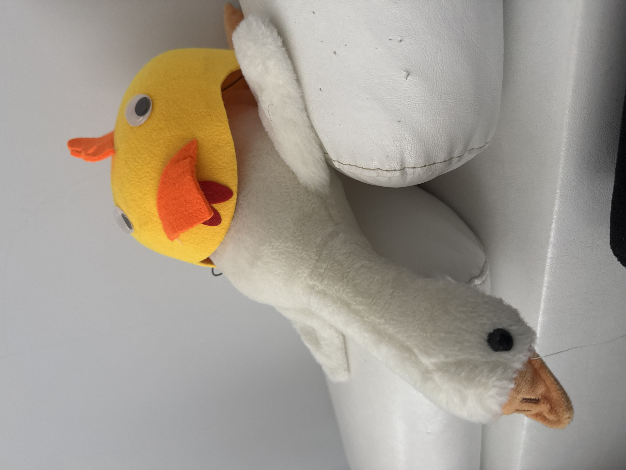 A plush white goose toy with a yellow chick hat sits atop a white leather-like surface. The goose has a long neck, a small black eye, and an orange beak. The chick hat is yellow with orange wings and googly eyes. The background is a light gray wall. The location is not discernible from the image.