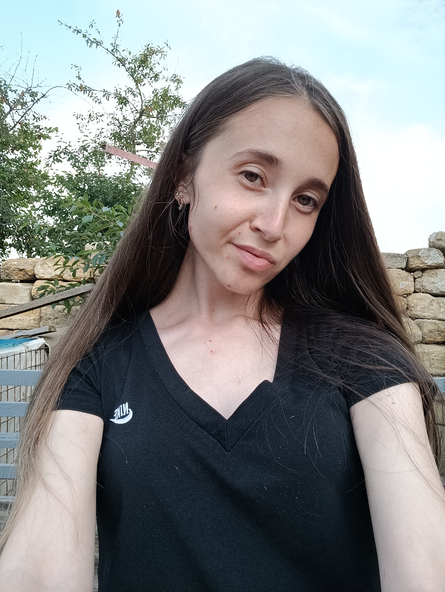 A young woman with long, dark hair is captured in a selfie outdoors. She is wearing a black V-neck t-shirt with a white Nike logo on the left side of her chest. Her hair is styled to one side, revealing her left ear with a small earring. The background consists of a stone wall, lush green trees, and a cloudy sky, suggesting a rural or suburban setting. The lighting is natural, indicating it is daytime. The photograph appears to have been taken in the city of Vypasne, Ukraine, based on the provided context.