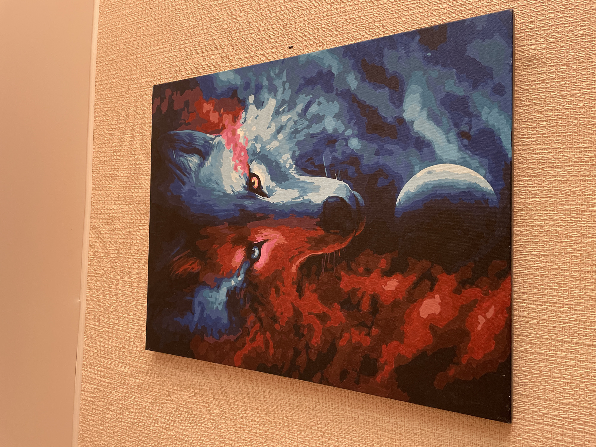 A painting of a wolf's face, bisected by contrasting colors, hangs on a textured, peach-colored wall. The left side of the wolf's face is rendered in vibrant reds and pinks, with a bright blue eye. The right side of the face is depicted in blues and purples, with a golden-orange eye. A crescent moon is positioned below the wolf's face, on the blue side. The painting is framed by a thin black border. The wall it is hanging on has a subtle horizontal line across the top, possibly indicating the edge of the ceiling. The lighting in the image suggests an indoor setting. There is no indication of the city of Laishevo, Russia, within the image.
