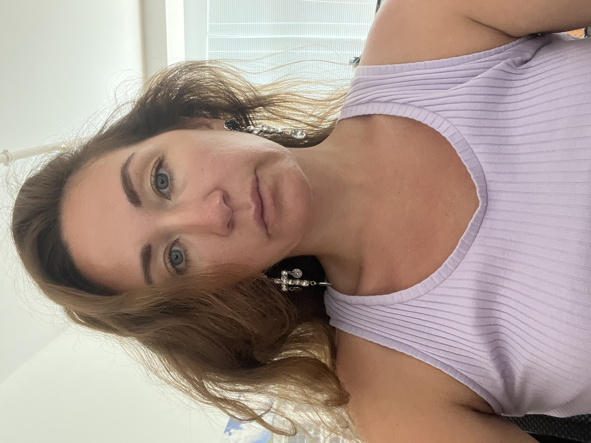 The image shows a close-up, chest-up shot of a woman with medium-length, wavy, brown hair. She has blue eyes and is wearing a light purple ribbed tank top and large, dangling earrings with cross-shaped charms. The background is out of focus and appears to be an indoor setting with blinds visible on the right. There is no indication of the city of Sochi, Russia in the image.