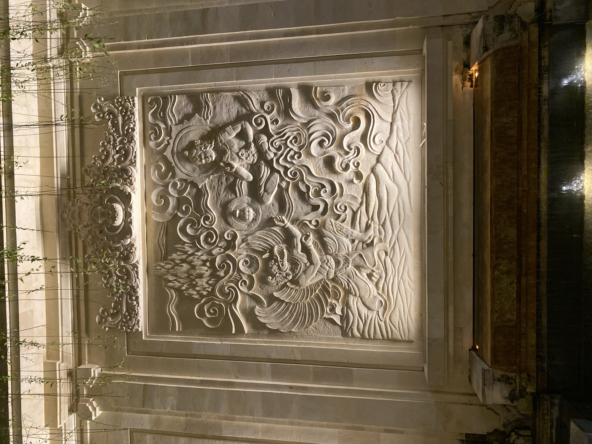 This image showcases an intricately carved stone relief panel mounted on a building facade. The relief depicts mythological figures and swirling cloud-like patterns, rendered in a pale, light-colored stone. The style suggests an Asian influence, possibly Balinese given the location context of Kuta Selatan, Indonesia. On the left side of the panel, a smaller, decorative carved element with floral motifs is visible. The scene is illuminated by artificial lighting, suggesting it is nighttime. The light casts shadows that accentuate the depth and detail of the carving. To the right of the relief, a vertical water feature or a dark, reflective surface is present, with water cascading down, also illuminated and creating a dynamic visual contrast with the static stone carving. The overall impression is one of artistic craftsmanship and atmospheric nighttime illumination.