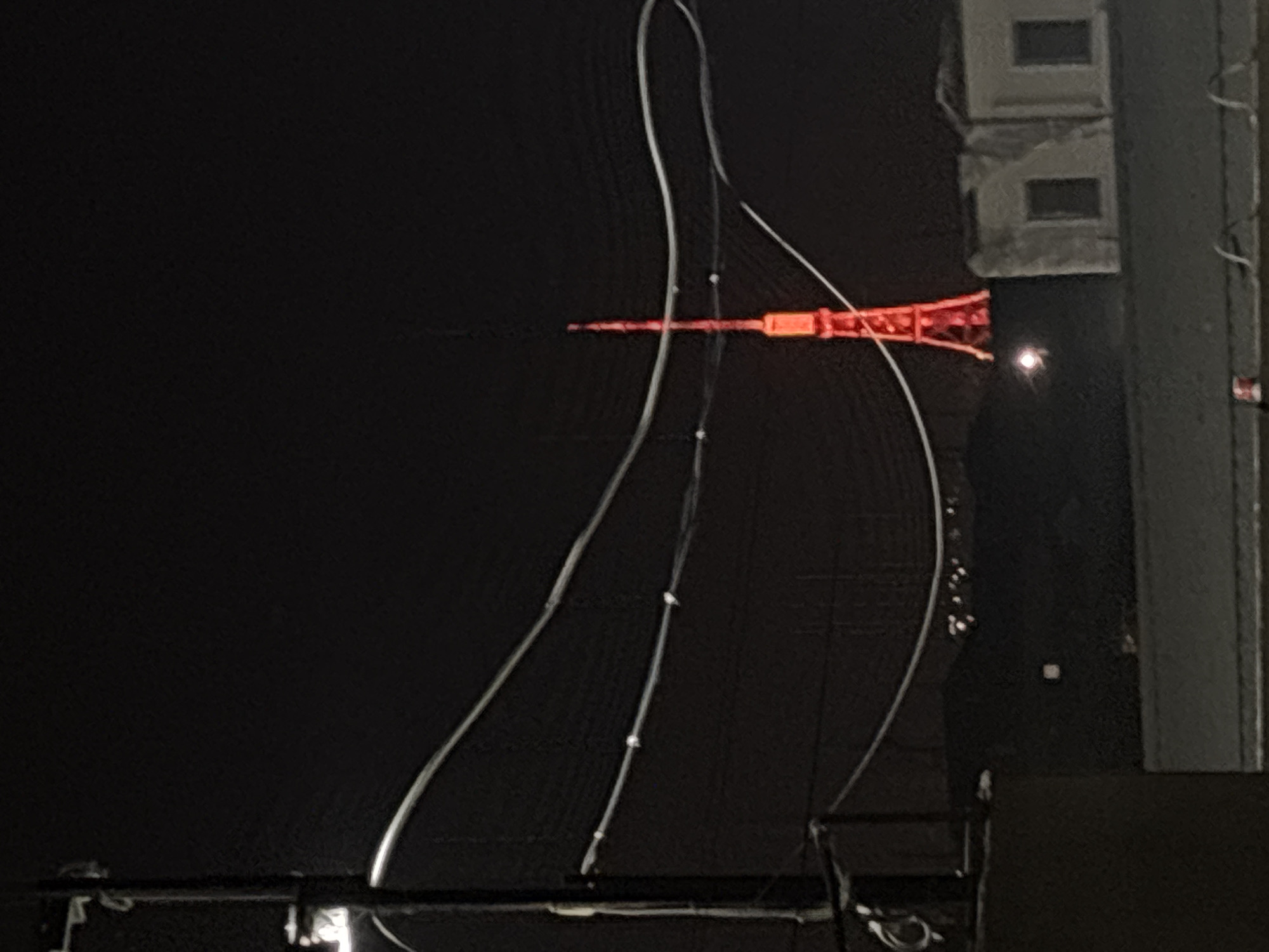 The image shows the Yerevan TV Tower illuminated in red at night. A thin, curved wire or cable is visible in the foreground, obscuring part of the tower. To the right of the tower, there is a dark building with illuminated windows. The surrounding environment appears to be dark, suggesting it is nighttime.