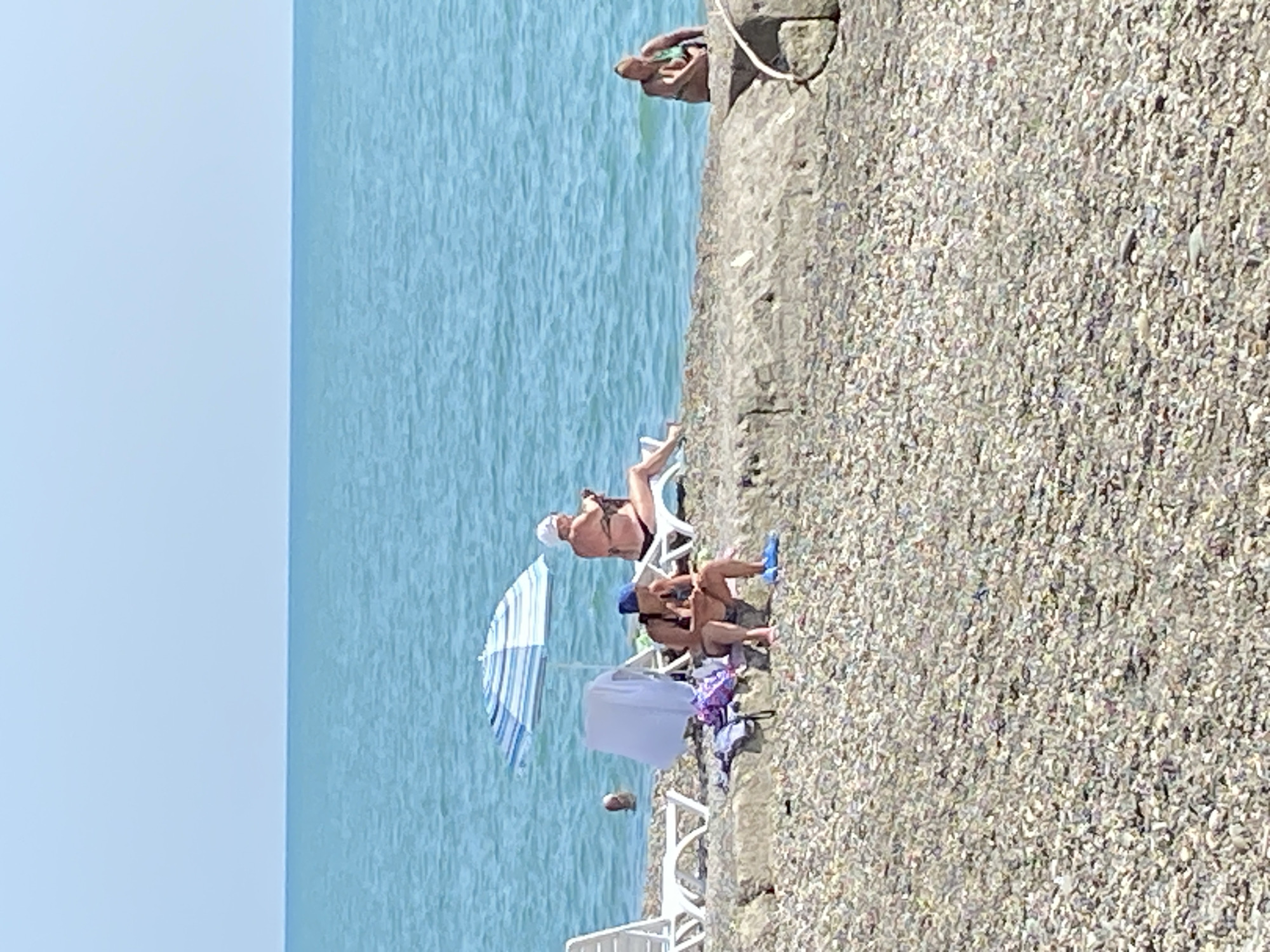 This is a medium-distance shot of a pebble beach in Chabkwara, Abkhazia, with the Black Sea stretching out to the horizon under a clear sky. In the foreground, a rocky, pebbled embankment forms the shore.
Several people are present on the embankment. On the left, two individuals are seated on the embankment. One, wearing a blue cap, is looking down at something in their lap, while the other, in a dark swimsuit and a blue cap, is also looking down. To the left of these individuals are two white beach chairs and a blue and white striped beach umbrella, with a white towel draped over one of the chairs. Further out in the water, a person's head is visible.
To the right, a woman in a black bikini is reclined on a white beach chair, looking down at something in her hands. Further to the right and slightly submerged in the water, another person is sitting and partially visible. The water itself is a calm, turquoise blue.