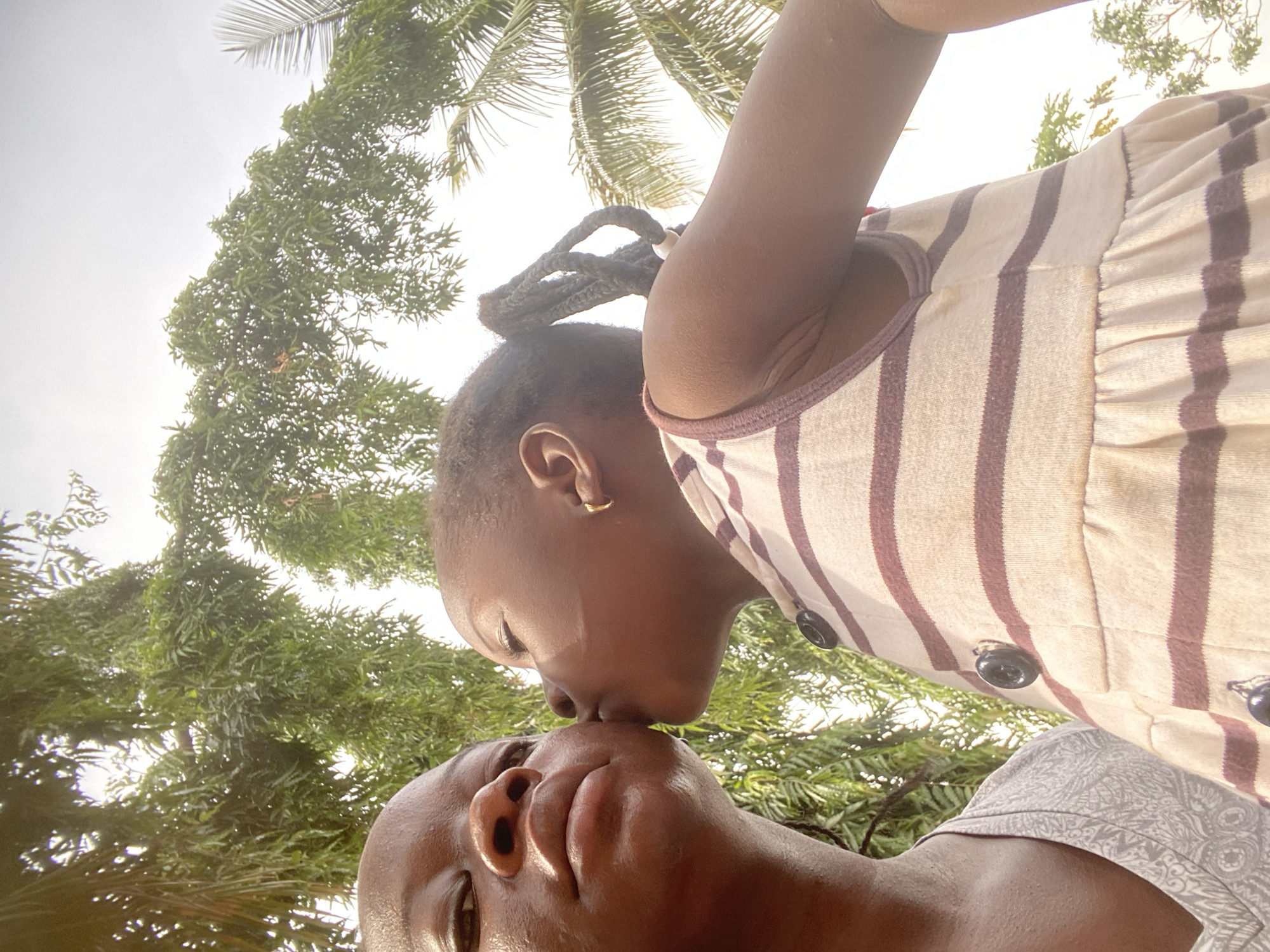 A low-angle shot features two dark-skinned individuals, presumably a parent and child, nose-to-nose under a lush canopy. The child, wearing a striped top with dark buttons, has braided hair and a small gold earring. Palm fronds and green foliage fill the background. The location is implied to be Maigizo, Nigeria.