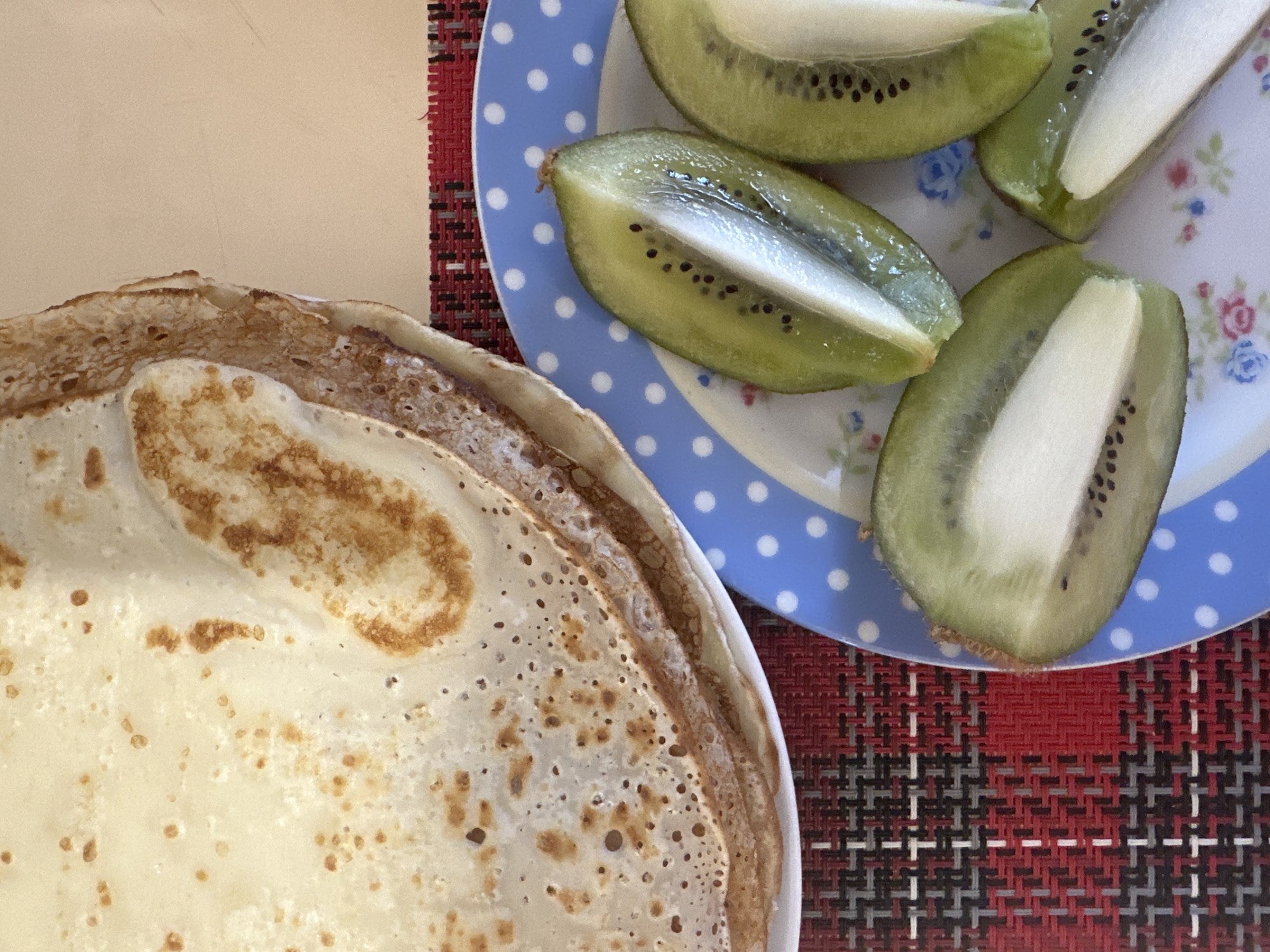 The image shows a close-up of a meal consisting of pancakes and kiwi slices. The pancakes are stacked on a white plate, showing a light brown speckled pattern. Adjacent to the pancakes, there is a plate with four slices of peeled kiwi, cut to reveal the green flesh, black seeds, and white core. The kiwi plate is blue with white polka dots and small floral designs. The plates are placed on a red, gray, and black checkered mat, which appears to be on a table, with a section of a tan surface visible in the upper left corner. No people are visible in the scene. The specific location in Votkinsk, Russia, cannot be determined from the image alone.
