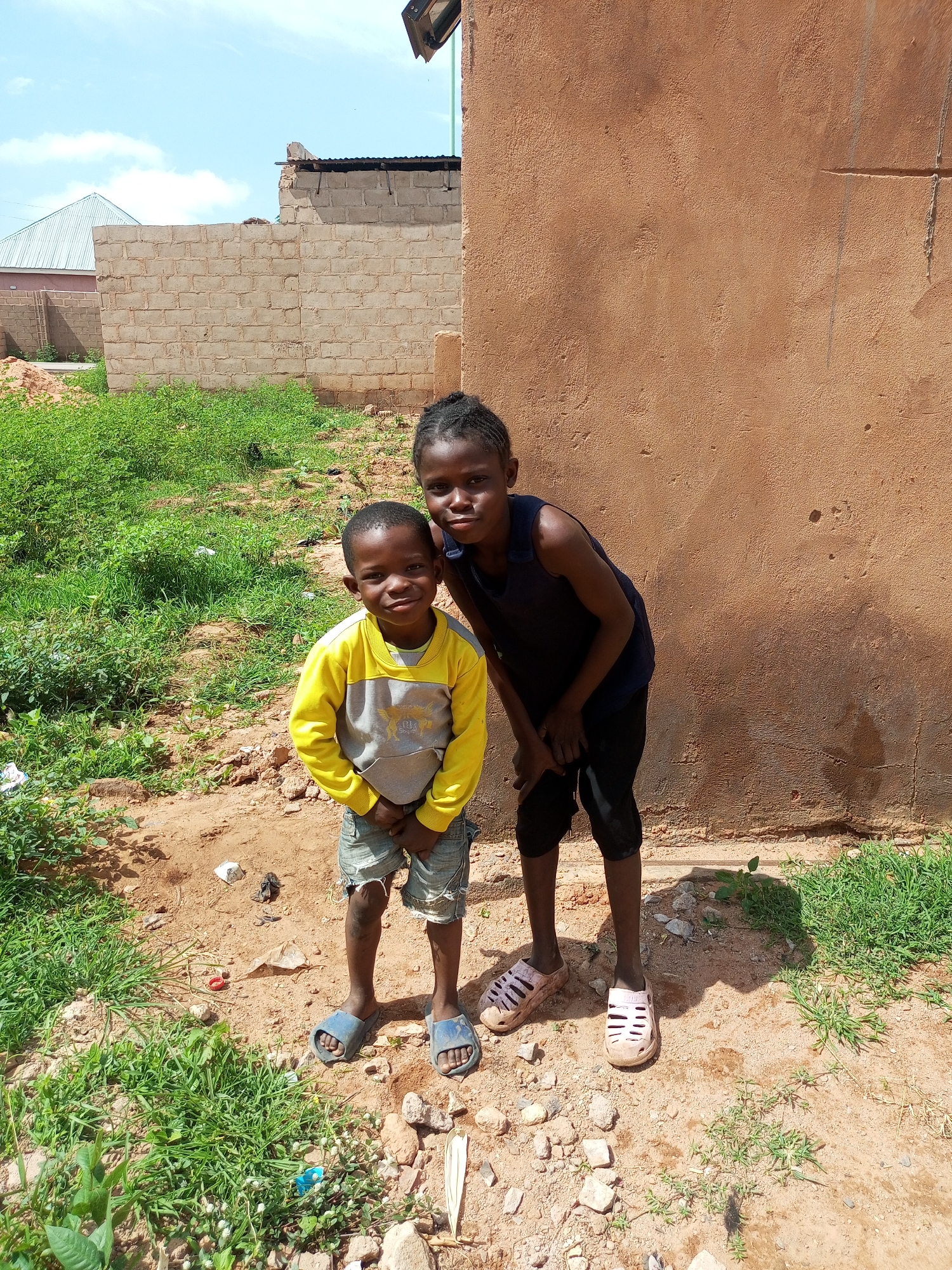 Two children stand in front of a rust-colored building wall, with a brick wall and vegetation in the background. The child on the left, a boy, is wearing a yellow and gray long-sleeved shirt, denim shorts, and blue sandals. The child on the right, a girl, is wearing a dark blue sleeveless top and black pants, with pink sandals. The ground is a mixture of dirt, small rocks, and patches of green grass. The sky above is bright and cloudy. The scene appears to be in an outdoor setting in Tempure, Nigeria.