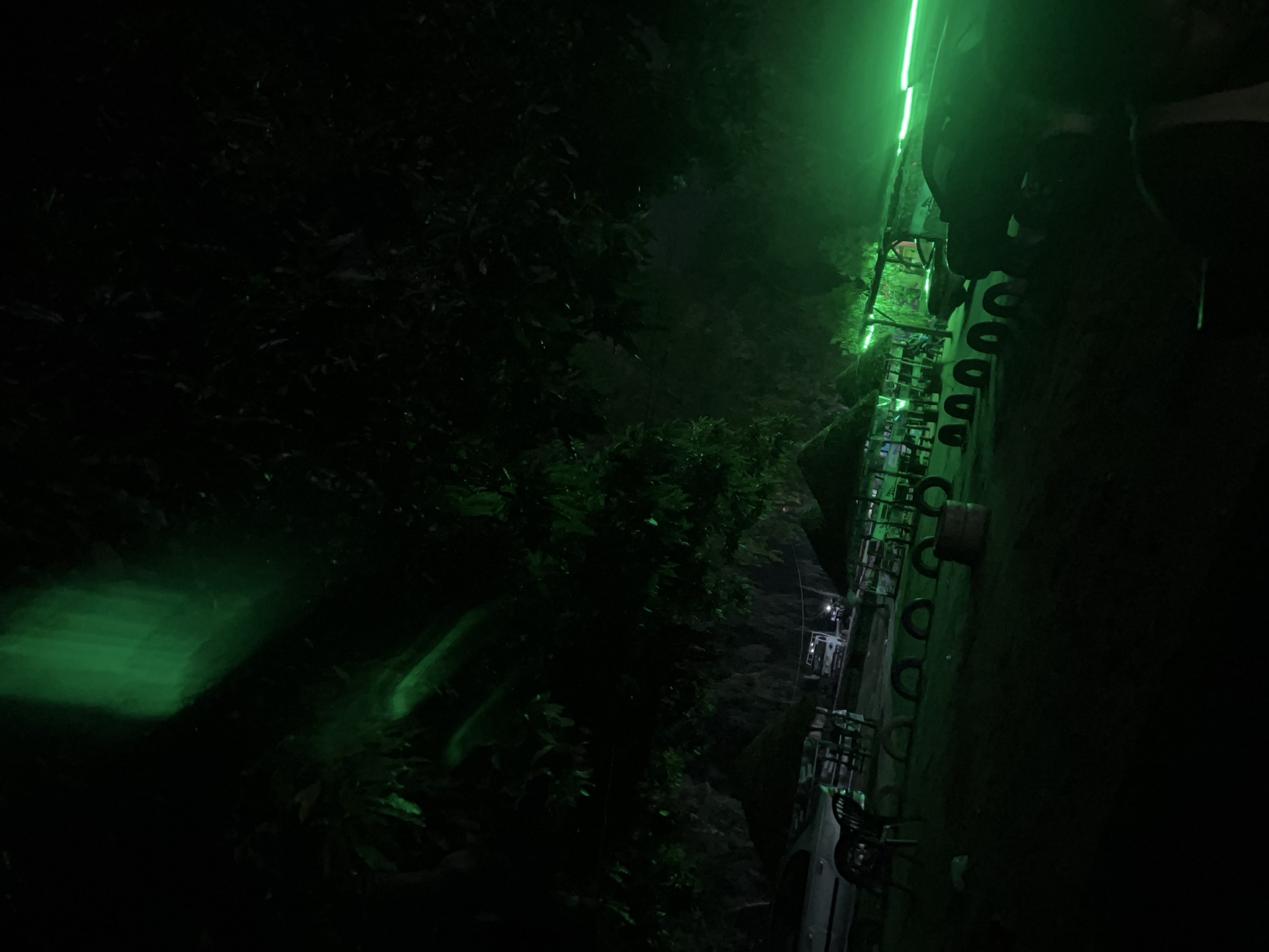 The image depicts a nighttime scene in a dimly lit outdoor area, possibly a recreational space or a rest stop, located in Howayi, Nigeria. The environment is characterized by dark foliage overhead, with prominent streaks of green light illuminating parts of the trees and the general atmosphere. In the foreground, several car tires are arranged on the ground, suggesting a rustic or informal setup. A few small thatched-roof structures, possibly shelters or seating areas, are visible further back. A white car is parked on the left side of the image. Beyond that, a white truck is also visible. The lighting, dominated by a vibrant green glow, creates a somewhat surreal or atmospheric effect. There are no clearly discernible individuals in the frame. The scene appears tranquil and quiet, with the green lights being the most dynamic element.