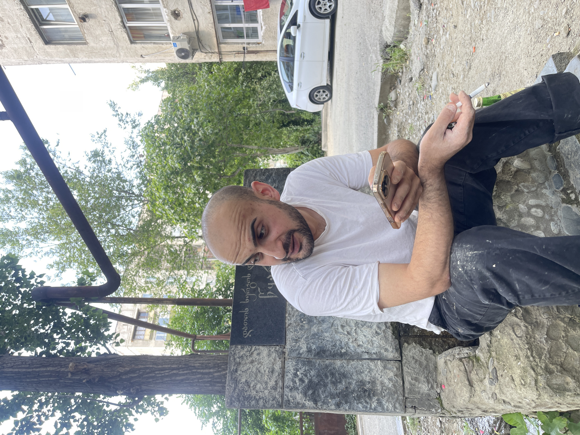 A bald, bearded man wearing a white t-shirt and black pants sits on a stone structure in Tbilisi, Georgia. He holds a gold-colored smartphone in his left hand and a cigarette in his right hand. A green bottle and a cigarette butt are on the ground near his right leg. In the background, a white car is parked on a street next to an apartment building with an air conditioning unit. Trees and greenery surround the man and the building. A stone monument with inscriptions is visible behind the man.