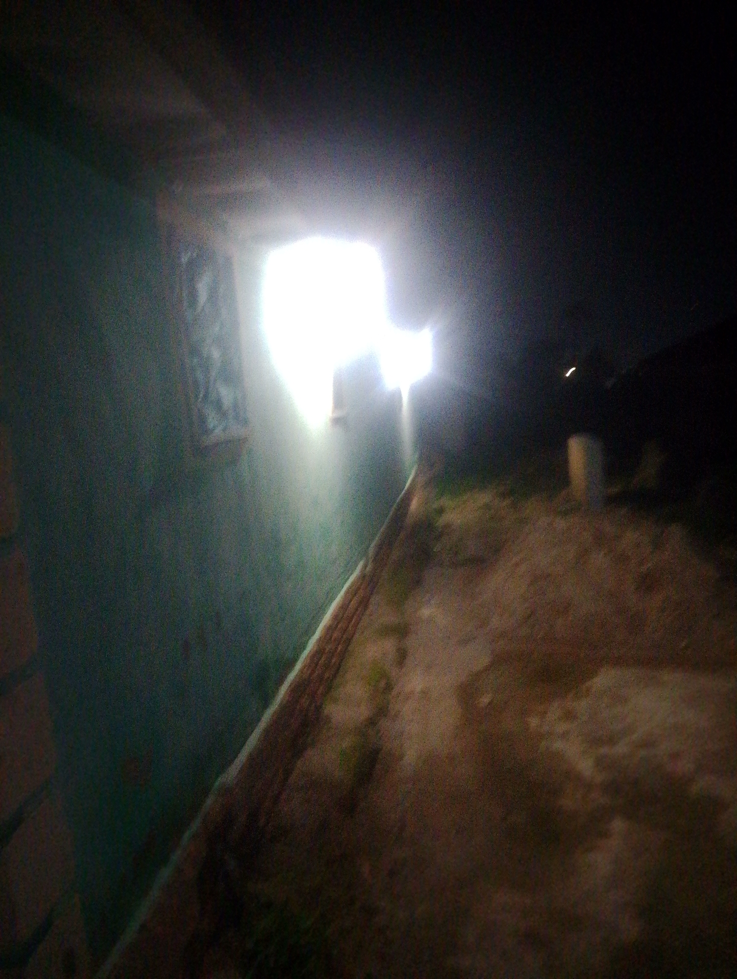 The image depicts a dark, outdoor scene with a bright light source emanating from an opening in a structure. A greenish-colored wall occupies the left side of the frame, with a patterned rectangular object visible on it. A brick-like structure runs along the bottom edge of the wall. The ground in front of the structure appears to be dirt or gravel. In the background, a cylindrical object, possibly a barrel or post, is visible. The overall atmosphere is dark and somewhat desolate. It is not possible to determine the specific location within Jos, Nigeria, based on the visual information provided.