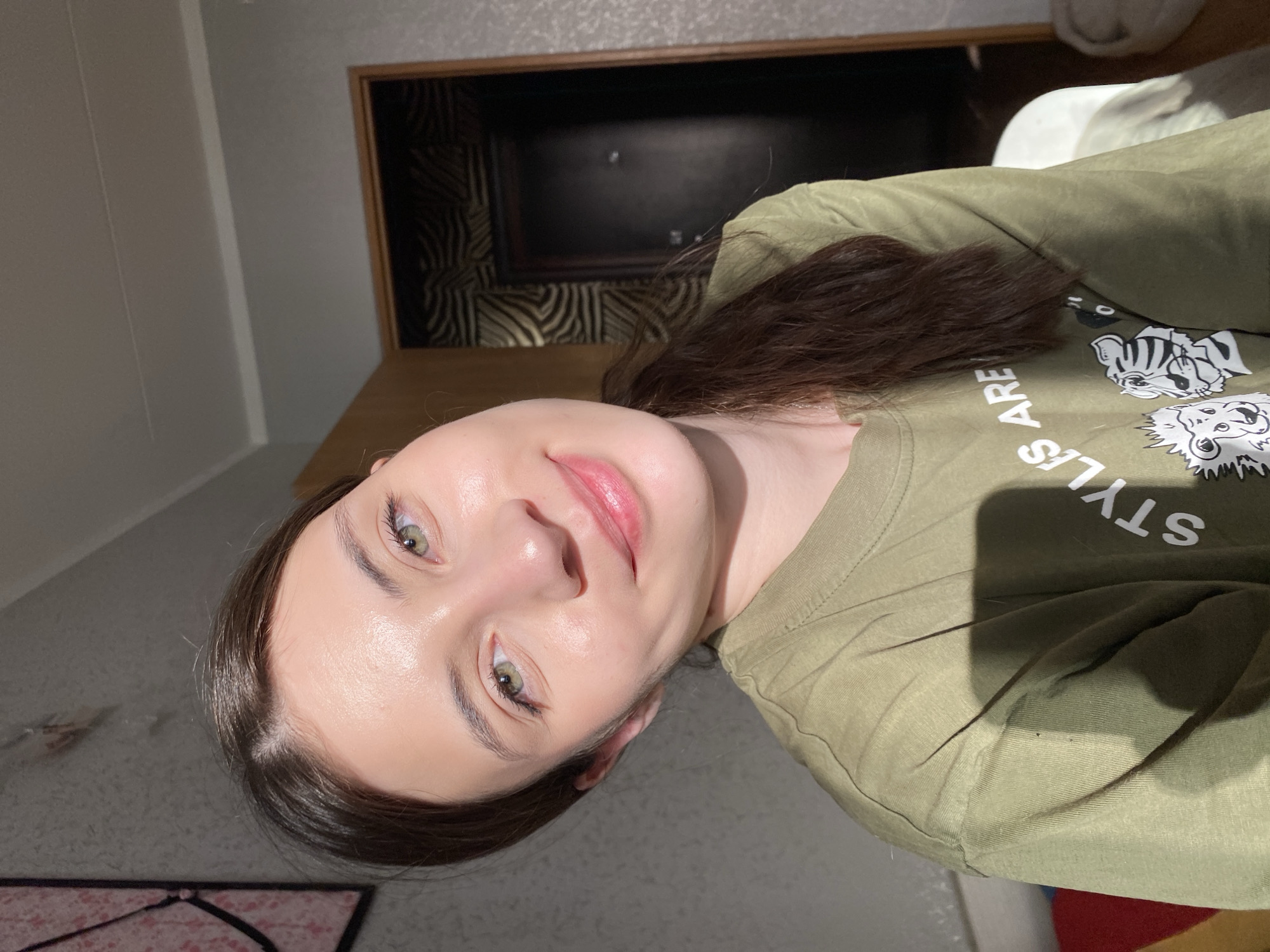A woman with brown hair and green eyes is pictured indoors. She is wearing an olive green t-shirt with a white graphic design. The background shows a bed or dresser made of wood with a patterned headboard. The location is potentially a residential setting in Votkinsk, Russia.