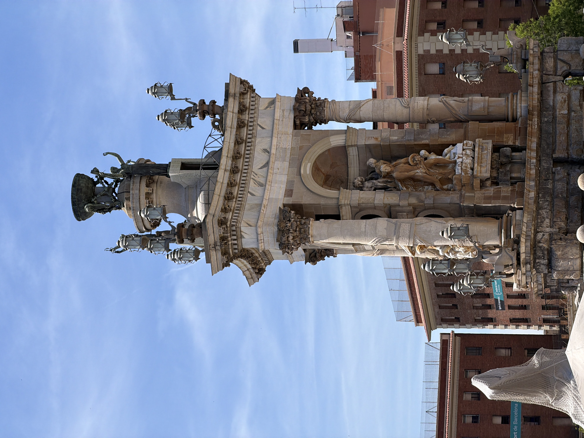 This image captures a detailed view of the Font de la Cascada, a large monument and fountain located in Barcelona, Spain. The monument is intricately designed with classical architecture, featuring multiple tiers, columns, and sculptures. On the upper tier, a bronze statue of Fame is visible, holding a laurel wreath, and above it sits a large bowl-like structure. Several ornate lanterns, also made of bronze, are strategically placed around the monument, some with multiple lights.
The central section of the fountain showcases a group of sculpted figures, appearing to depict mythological or allegorical scenes. The material used for the main structure is stone, with various shades of beige and grey, and there are decorative elements with floral motifs.
In the background, modern buildings with brick facades and tiled roofs are visible, indicating an urban setting. A bright blue sky with scattered white clouds forms the backdrop. On the left side of the image, part of another structure covered with a netting-like material is seen. The overall scene is well-lit, suggesting it was taken during daytime.