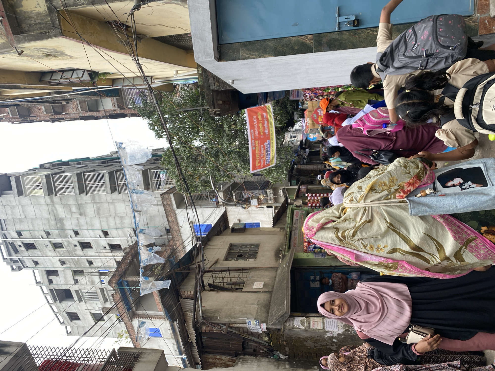 The image depicts a street scene in Dhaka, Bangladesh, characterized by dense urban development and bustling activity. Dominating the left side of the image is a tall, concrete building under construction, with exposed rebar and blue plastic sheeting. Older, dilapidated buildings with corrugated metal roofs and exposed brick are interspersed with the modern structure. Overhead, a tangled network of electrical wires crisscrosses the scene.
On the right side of the image, a group of people, primarily women and children, are gathered on the sidewalk. Many of the children are wearing school uniforms and carrying backpacks. A woman in the foreground is wearing a pink hijab and a black dress, carrying a small purse. Behind her, other women are dressed in traditional clothing. A banner advertising "ABACUS MATH" and "Sangeet Proshikhon Course" (music training course) in Bengali is visible, indicating the presence of an educational institution. The scene is crowded and suggests a vibrant, but somewhat chaotic urban environment. The overall impression is one of a densely populated area undergoing rapid development, with the juxtaposition of old and new structures reflecting the city's evolving landscape.