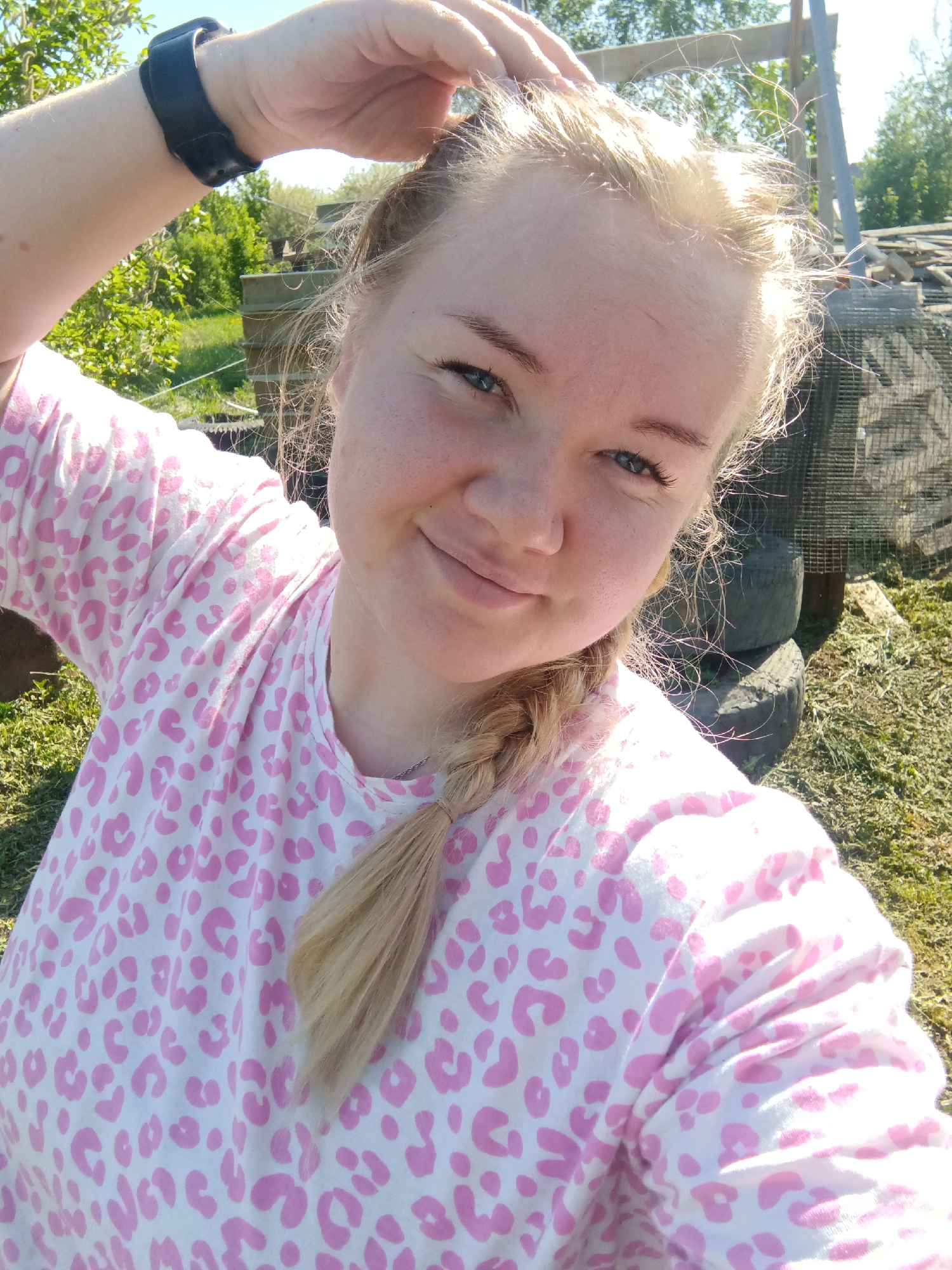 A woman poses for a selfie in an outdoor setting in Unknown, Unknown. She is fair-skinned with blonde hair styled in a braid, and she wears a pink and white leopard-print long-sleeved shirt. Her left hand is raised, appearing to touch her hair. A black wristwatch is visible on her left wrist.
Behind her is an untidy backyard area with overgrown grass. There are stacks of used tires visible near a wire mesh fence, along with other miscellaneous items and materials, possibly related to a construction or repair project. In the distance, trees and greenery suggest a more rural or suburban setting. Sunlight is present, indicating it is daytime.