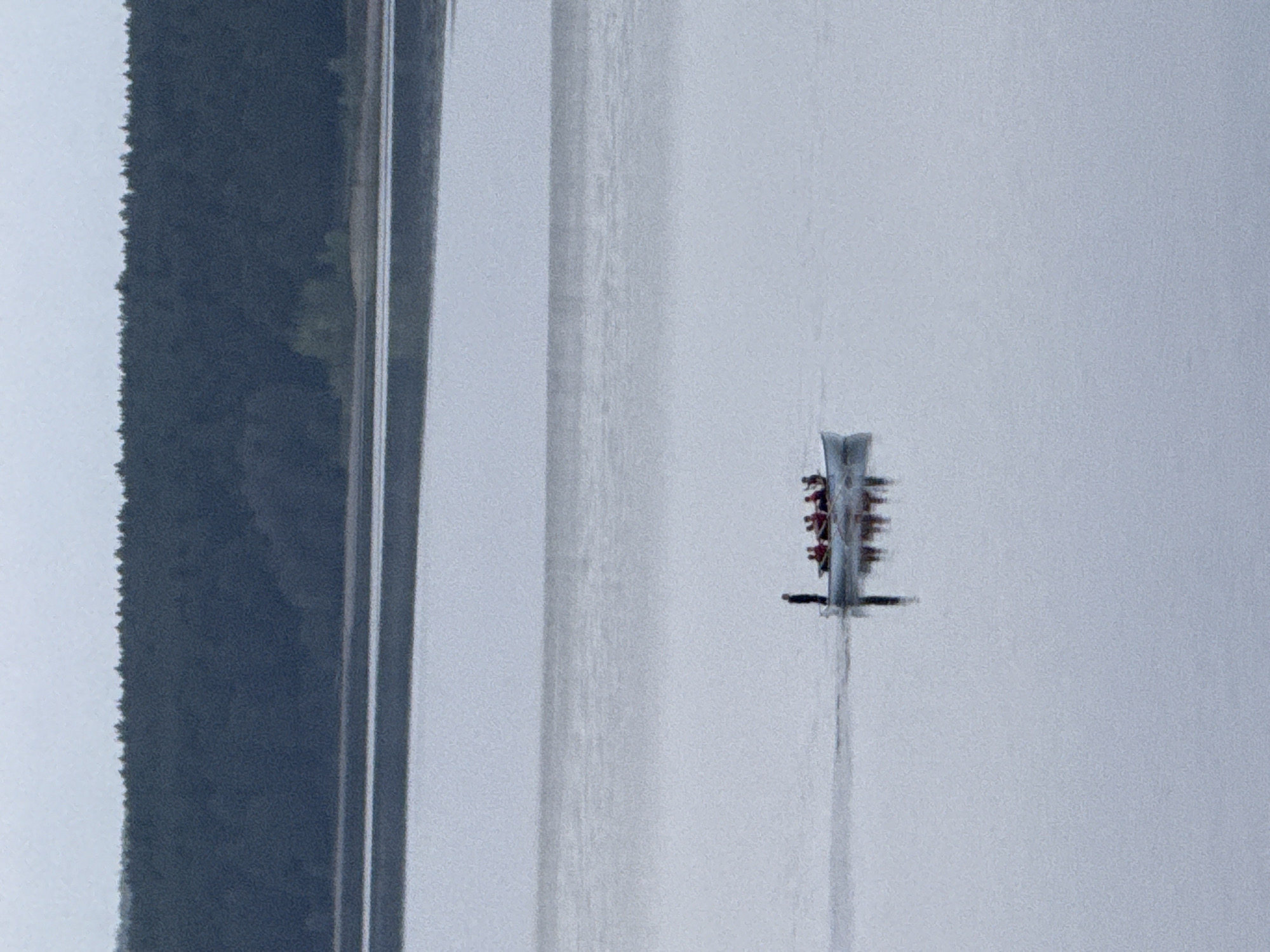An aerial view of an eight-person rowing crew in a boat on the Votkinsk Reservoir, Russia. The boat is moving on the calm water. The coastline with trees is on the left side of the image.