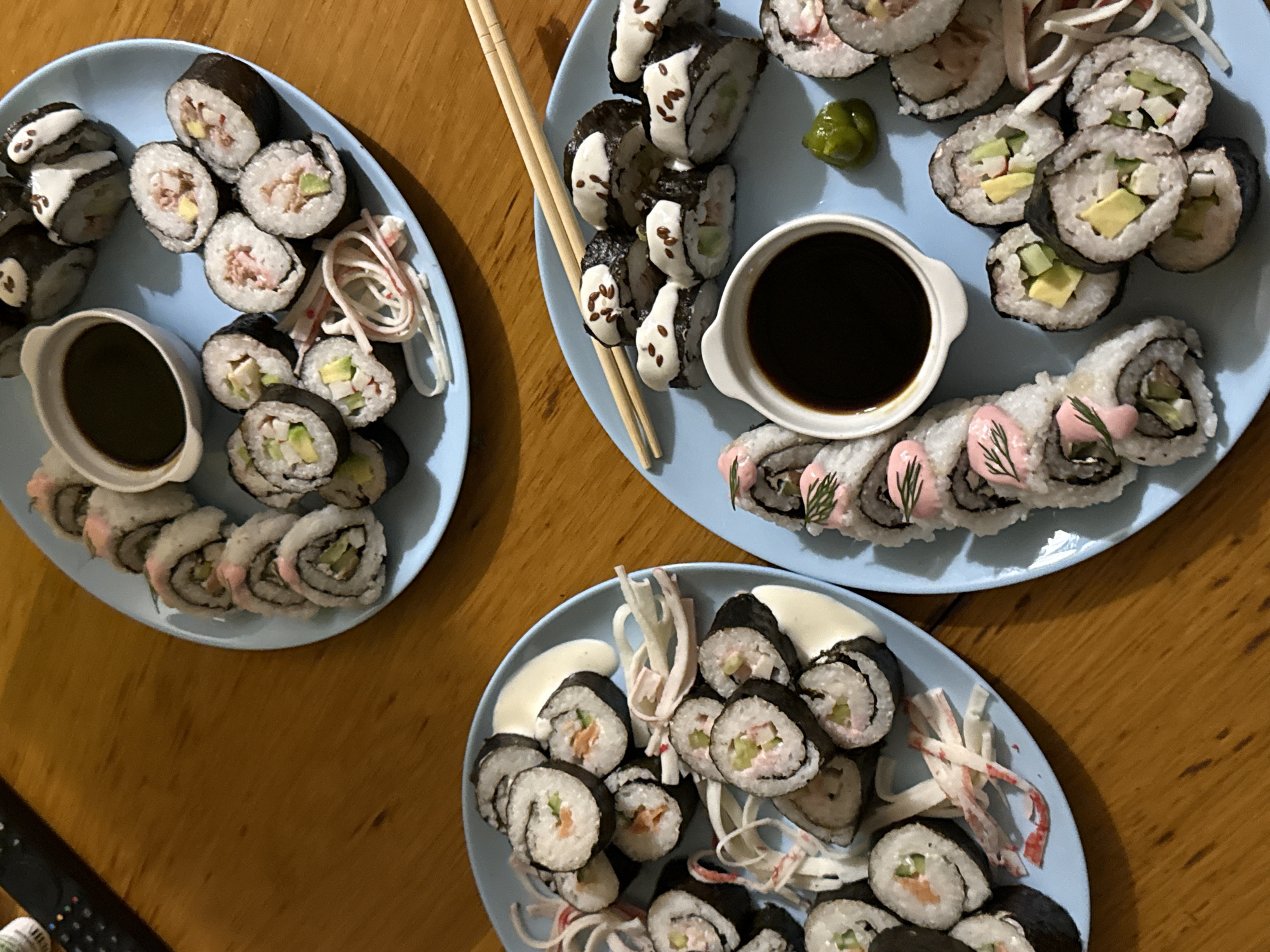 The image depicts a wooden table on which three light blue plates are arranged. Each plate contains a variety of sushi rolls. One plate contains sushi rolls with avocado and crab meat, as well as some decorated with white sauce and imitation crab sticks. Another plate includes sushi rolls decorated with white sauce and small black "eyes," alongside a small dish of soy sauce and chopsticks. The third plate features sushi rolls with avocado and tuna, as well as some decorated with pink sauce and what appear to be dill sprigs, along with a small dish of soy sauce. No people are visible in the image. The location is unknown, and there is no indication it is in Sofia, Bulgaria.