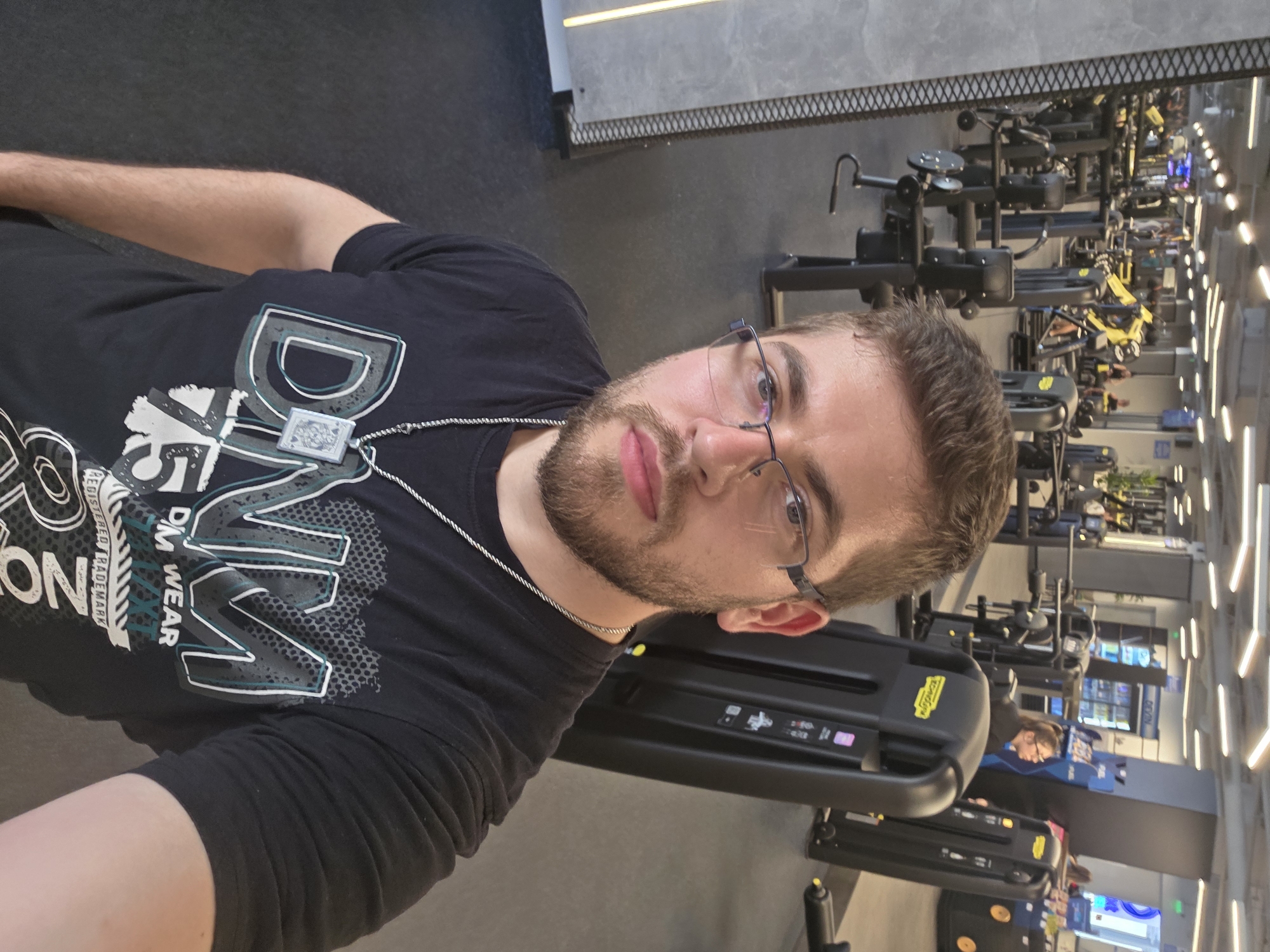The image shows a man wearing glasses, a black t-shirt with a design, and a necklace, inside a gym. The background shows rows of exercise machines and other gym equipment. Other people are present in the background, using the equipment. The lighting is bright and appears artificial. The location is likely a gym in Plovdiv, Bulgaria.
