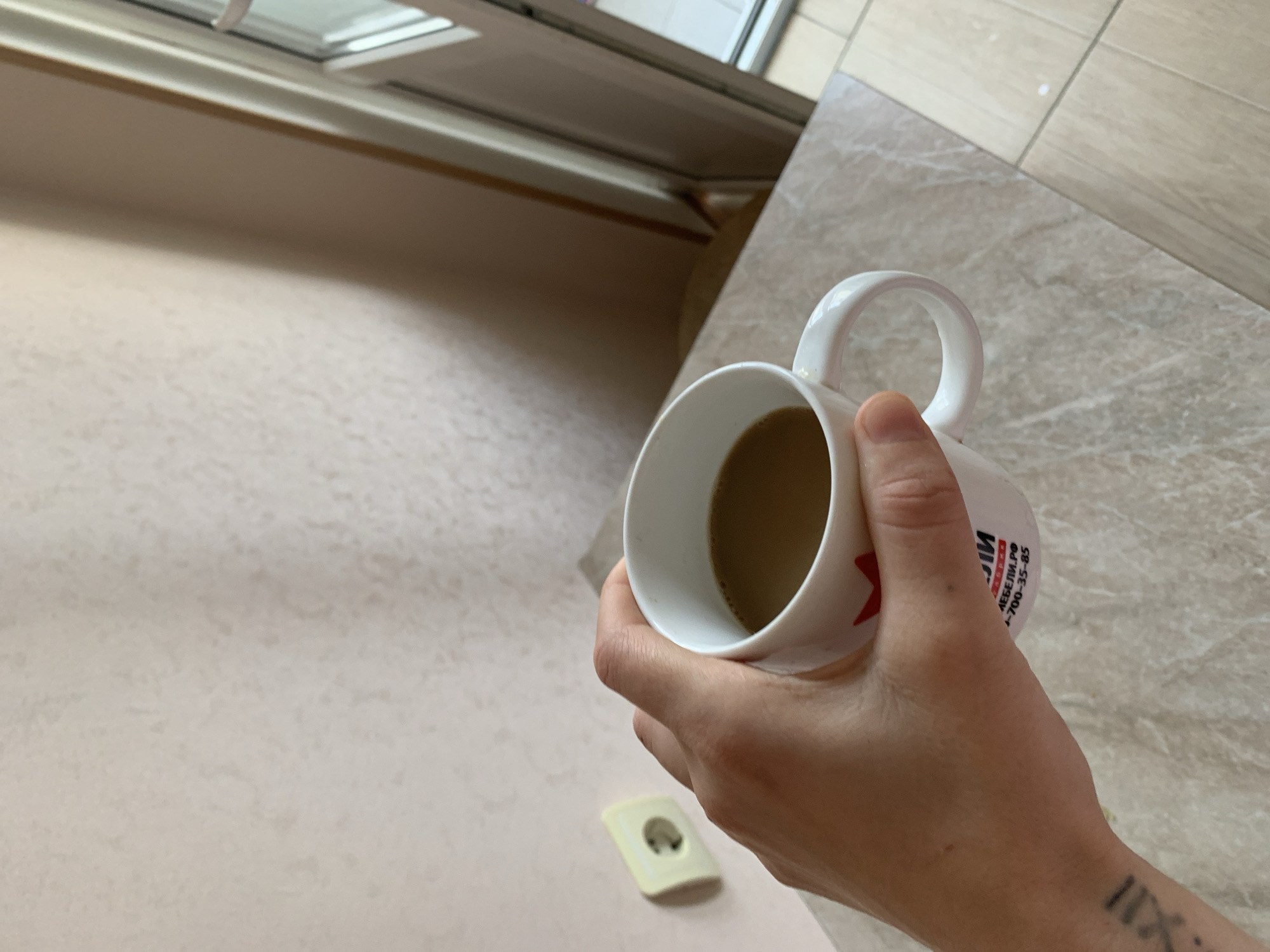 A person's hand holds a white ceramic mug filled with coffee. The mug has red lettering and a partial logo on its side. The background includes a beige textured wall with a single white electrical outlet and a tiled floor and countertop. A doorway or window with a white frame is visible to the right. A tattoo of Roman numerals is visible on the person's forearm.