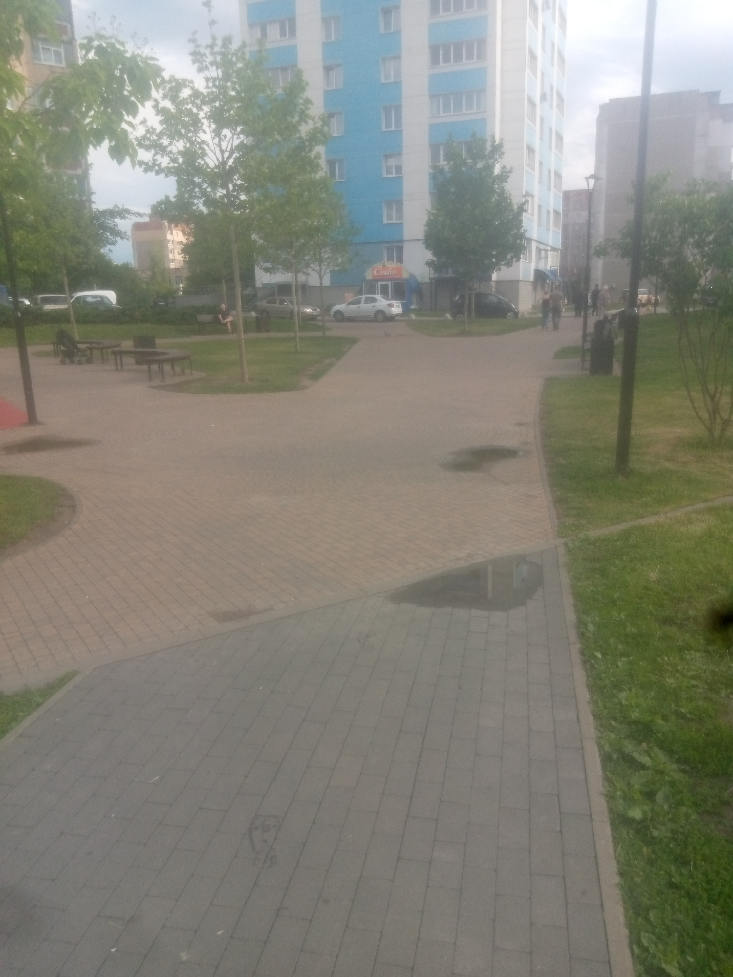 An outdoor scene in a residential area depicts a paved walkway with benches and tables, surrounded by trees and grass. In the background, a tall apartment building with blue and white accents stands prominently. Several cars are parked in the vicinity, and a few people are visible walking along the paths or sitting on a bench. The sky appears overcast.