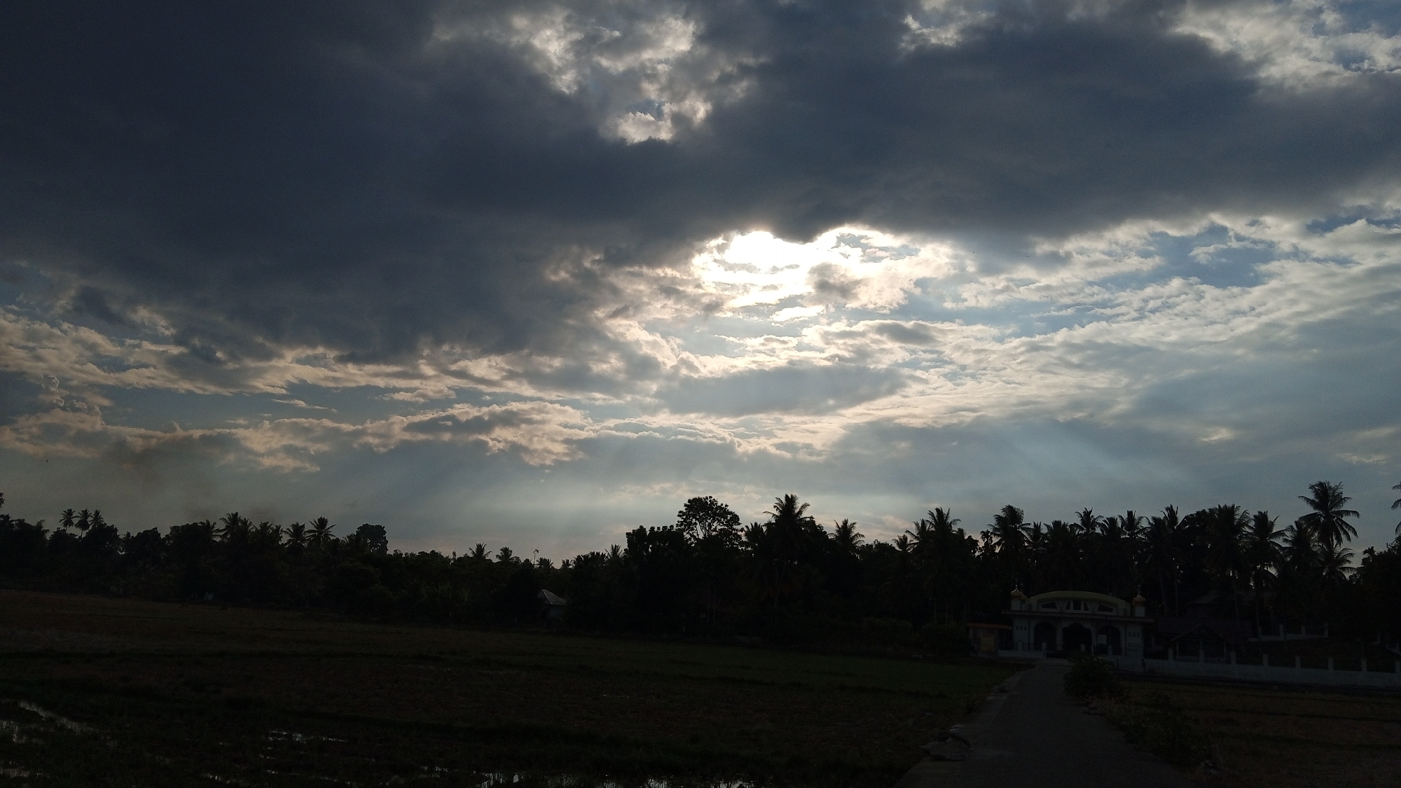 The image depicts a rural landscape in Blangreuling, Indonesia, under a dramatic sky filled with dark and light clouds. Rays of sunlight pierce through an opening in the clouds, creating a spotlight effect over a treeline. The foreground is dominated by a dark, silhouetted agricultural field, possibly rice paddies with standing water. To the right, a paved road leads towards a building with a distinct arched entrance, likely a mosque or a similar structure, adorned with yellow and white accents. Coconut palms and other trees form a dense silhouette along the horizon, with a hint of smoke rising from the left side of the treeline, suggesting a distant fire or cooking activity. The overall atmosphere is one of natural beauty and rural tranquility, with the dramatic sky adding a sense of awe. No people are visible in the frame.
