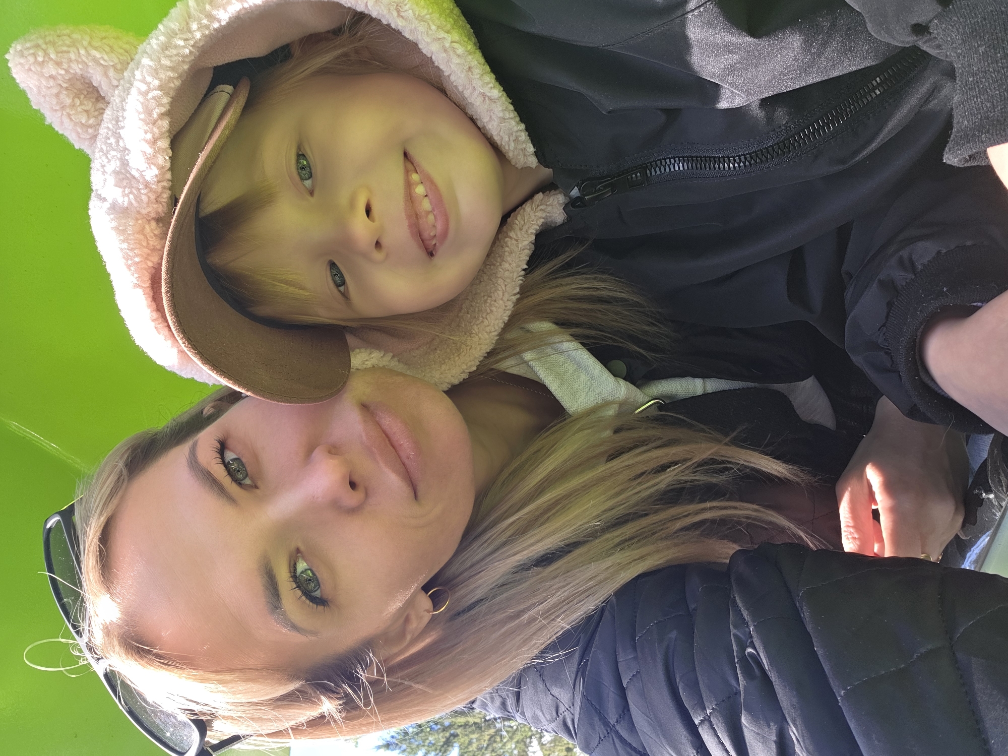 A woman and a young child, possibly mother and daughter, are posing closely for a selfie in what appears to be an outdoor setting. The background is predominantly green, possibly suggesting a park or grassy area.
The child is wearing a hooded jacket, the hood lined with a soft, light-colored material, possibly fleece or faux fur. The jacket also has a brown brim. The child is smiling, revealing their upper teeth, and appears to be looking directly at the camera.
The woman has light hair and is wearing a dark jacket. She is also looking at the camera. Sunglasses are visible in her hair.
The location is not discernible from the image. No details suggest that this image was taken in Ufa, Russia.