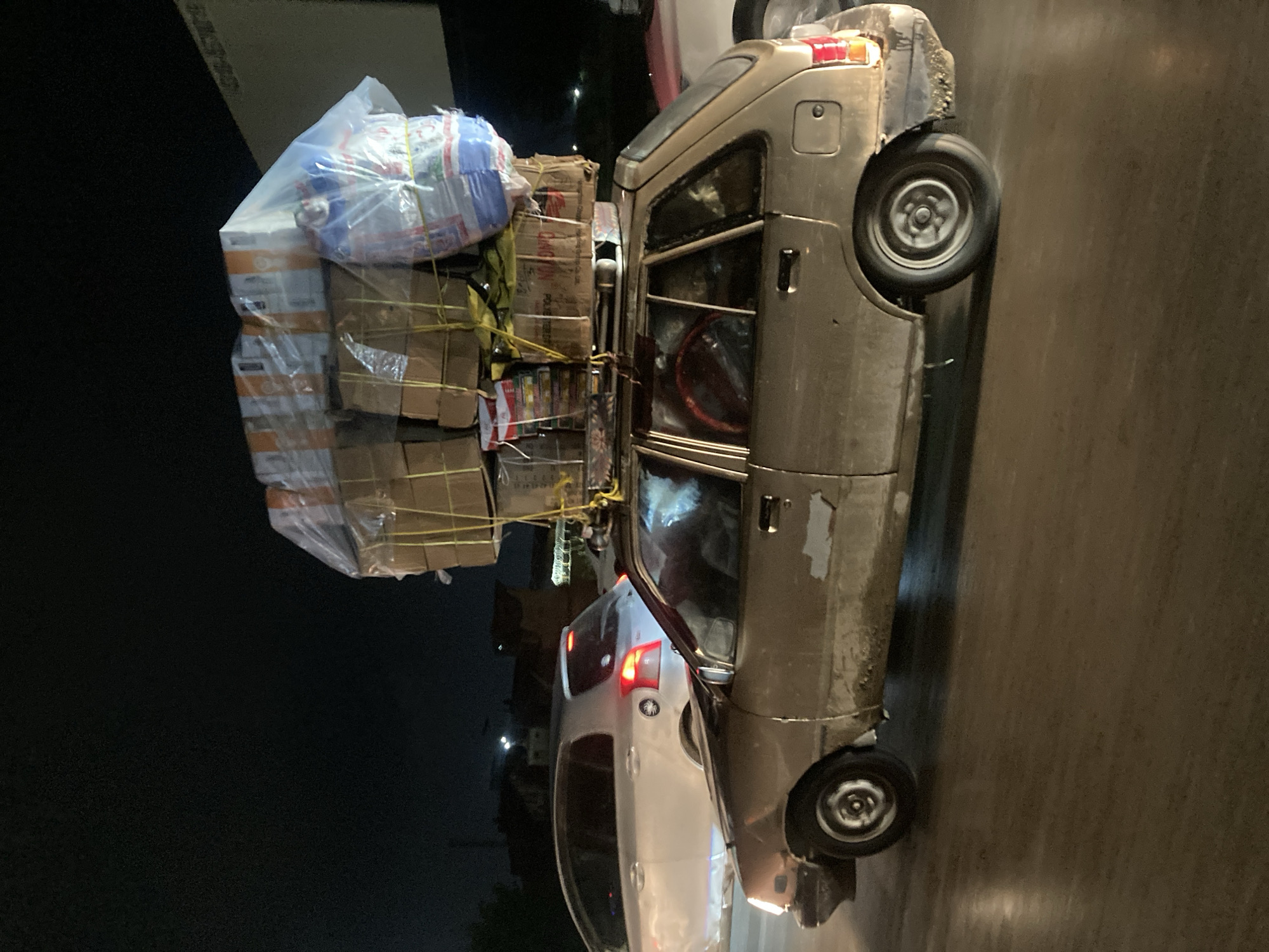 The image shows a light-colored vehicle heavily loaded with packages and plastic-wrapped items strapped to its roof. The vehicle appears to be in a city environment, possibly Quetta City Tehsil, Pakistan, as suggested by the context. The load is secured with rope. A white car is parked nearby. The scene is illuminated by artificial light, suggesting it is nighttime. The vehicle shows signs of wear and tear, indicating it is likely used for commercial transport.