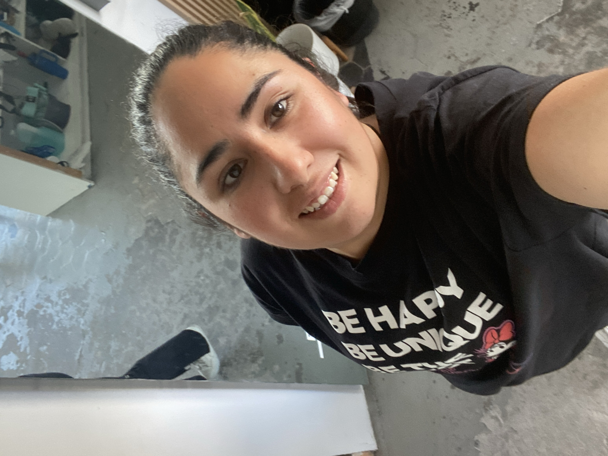 A woman with dark hair pulled back in a ponytail smiles into the camera while wearing a black t-shirt with white text and a cartoon image. The text on the shirt reads "BE HAPPY, BE UNIQUE." She appears to be standing in a room with a gray concrete floor and a white shelf or countertop visible on the left. The location cannot be determined from the information in this image.