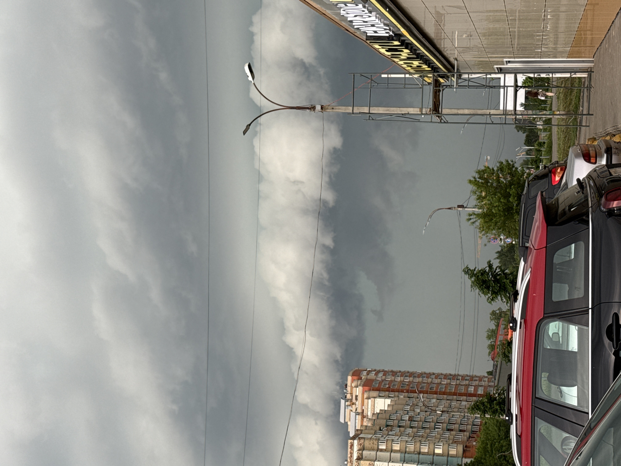The image shows an outdoor scene in Izhevsk, Russia, on a cloudy, overcast day. The sky is predominantly gray with heavy clouds. In the foreground, there are parked cars, including a red SUV, along a street. A tall, apartment building is visible with rows of windows, suggesting a densely populated urban area. There are trees and greenery interspersed with the buildings and cars, suggesting an urban setting. Street lights with curved arms are present, and scaffolding is attached to a building with signage in a language other than English. The overall impression is of a city street during an inclement weather.