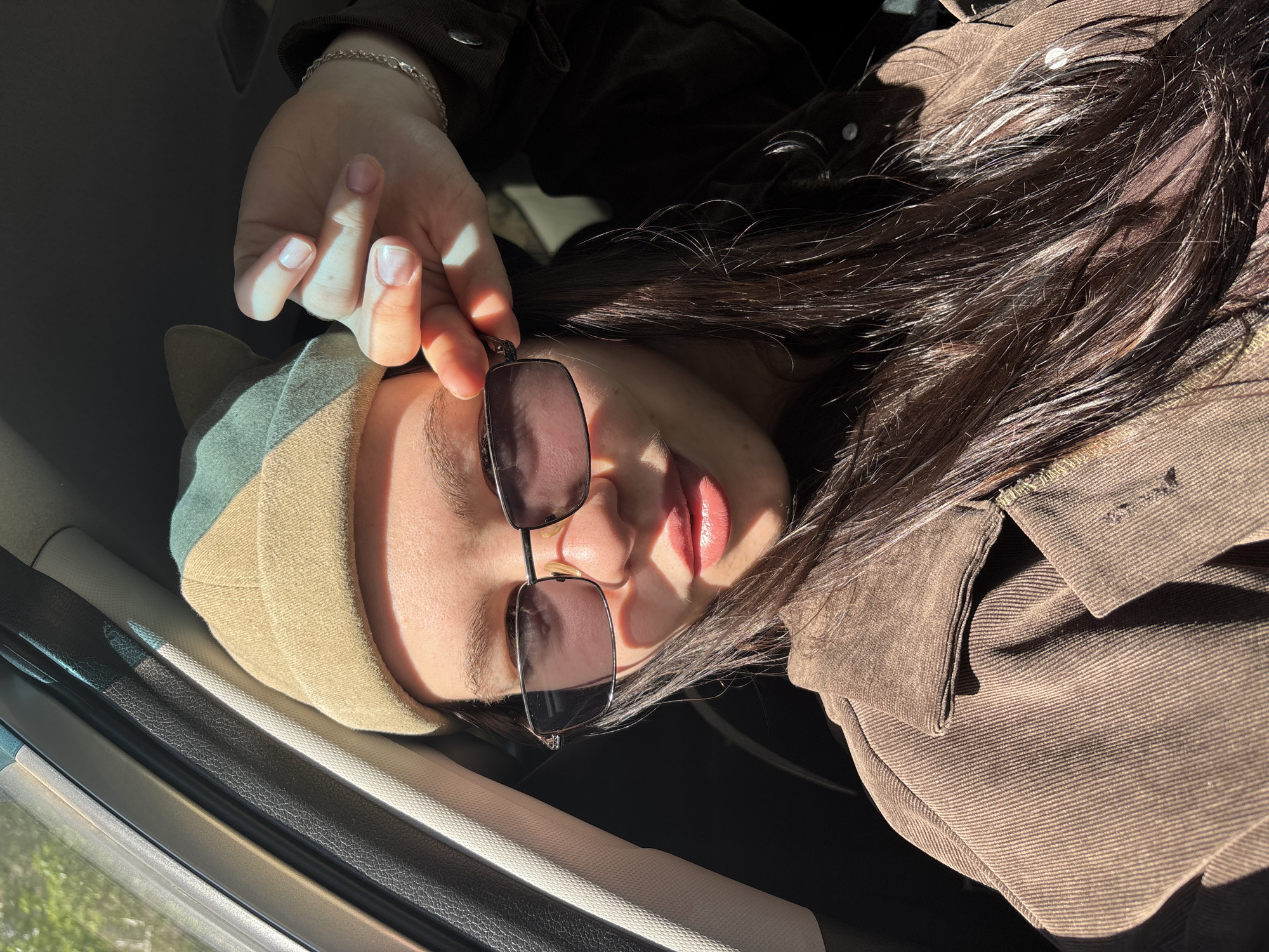 A young woman lies in a car, wearing a brown corduroy jacket and a khaki-colored beanie. She is holding up her sunglasses with her left hand. The scene is lit by strong sunlight. The location cannot be determined to be Perm, Russia from the image alone.