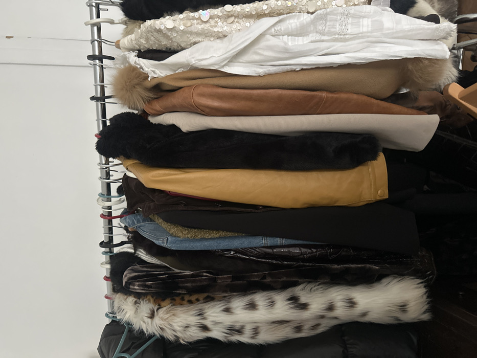 The scene depicts an indoor clothing display in Paris, France. A horizontal, chrome-finished metal clothing rack extends across the mid-ground, holding a collection of approximately fifteen assorted garments on various hangers. From left to right, the items include a black puffer coat, a white and brown spotted faux fur vest, a dark brown velvet coat, a dark textured jacket, a blue denim jacket, a black blazer, a mustard yellow leather jacket, a black faux fur coat, a light beige ribbed top, a brown leather jacket, a tan coat with a fur pom-pom detail, a white textured shirt, and a cream sequined jacket. The background consists of a plain white wall. The lower foreground reveals a dark floor, possibly wood, with a partial view of a wooden hanger lying on it. No individuals are present in the image.