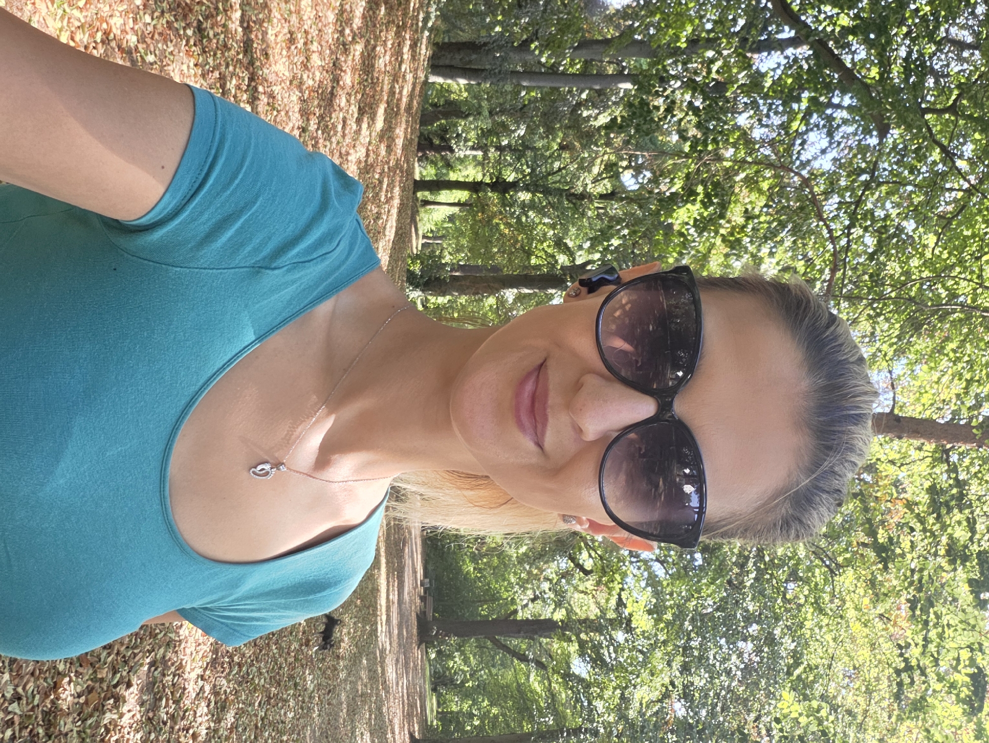 A blonde woman with sunglasses and a turquoise shirt is taking a selfie in a park. She is wearing earbuds and a necklace with a pendant shaped like baby footprints. The park has many trees with green leaves and fallen leaves on the ground. The sun is shining, casting shadows. In the background, a small dog is running. The location is likely a park in Munich, Germany, given the abundance of mature trees and well-maintained paths.