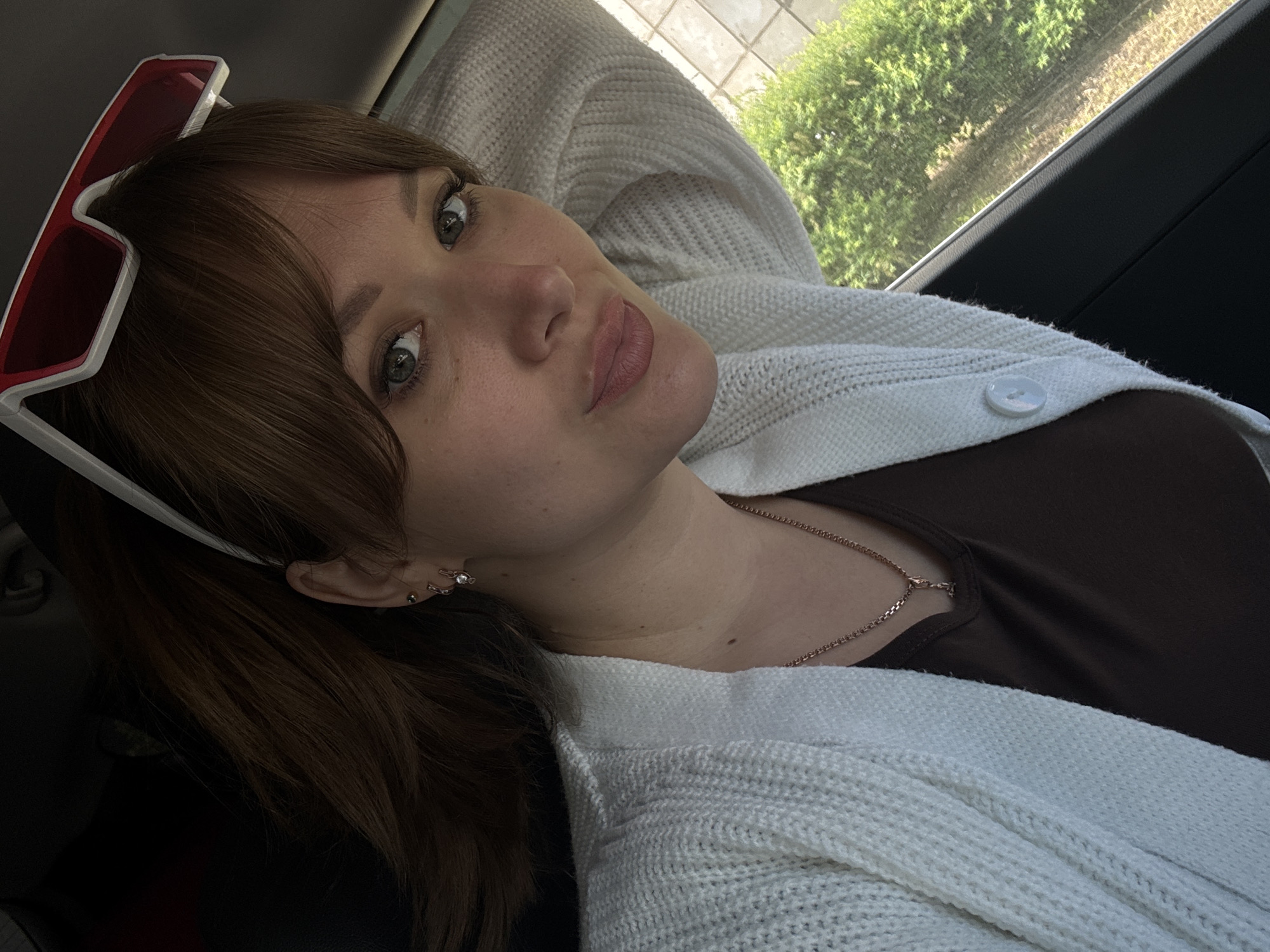 A close-up, angled selfie captures a young woman inside a car, looking directly at the camera with a slight pout. Her medium brown hair has bangs and she has light-colored eyes. Red and white framed sunglasses are perched on top of her head. Her visible ear features multiple small piercings. She is wearing a dark brown top beneath a loose-knit, white cardigan, fastened with a single white button. A delicate, silver-toned chain necklace is visible around her neck. Her right arm is partially seen, bent behind her head, suggesting a relaxed posture. Through the car window in the background, bright green foliage and sunlight are visible, indicating it is daytime. The scene suggests a casual moment, possibly while traveling. The location context for the image is Chaikovsky, Russia. There is no visible text in the image.