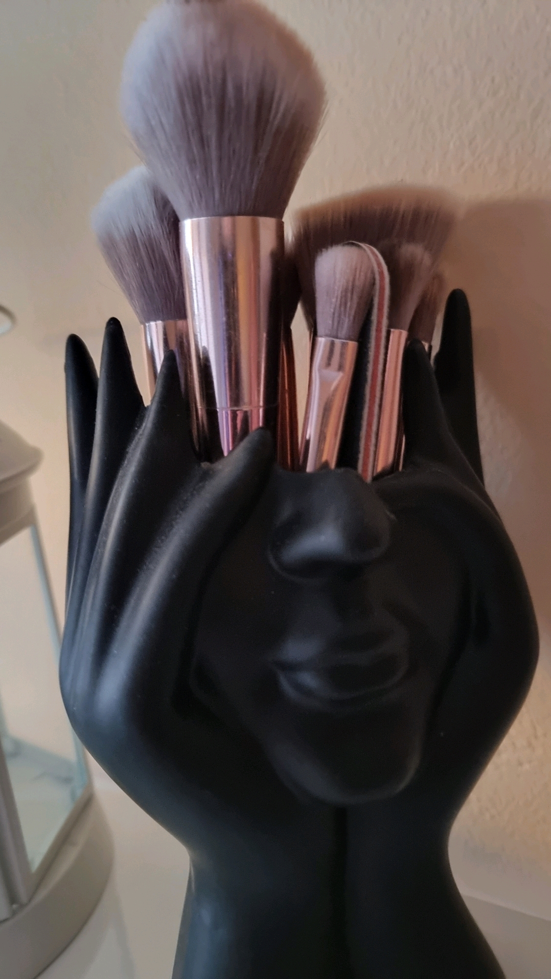 A black, abstract statue of a face with hands holding it is used as a makeup brush holder. Several makeup brushes with pink metallic handles and fluffy grey and brown bristles are stored in the holder. The background is a neutral-colored wall and a hint of a white shelf. The lighting is artificial and casts a soft shadow on the wall behind the statue. The statue's design suggests it is meant to evoke a feeling of support or artistry. No specific location in Sant Joan Les Fonts, Spain, is identifiable from the image.