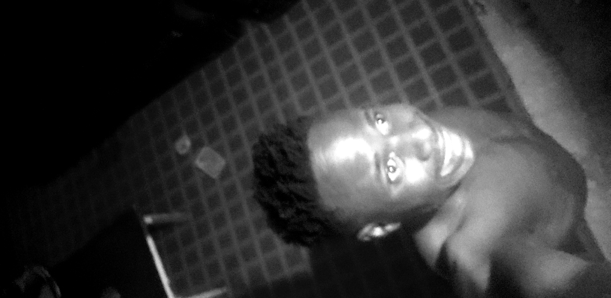 The image is a close-up, low-light, black and white, selfie-style photograph of a young Black male. He is shirtless and looking towards the camera with a wide smile, revealing his teeth. His eyes are looking slightly off-camera to his right. He has short, textured dark hair and visible ears. The background is dimly lit and shows a checkered pattern, suggesting a tiled floor or surface. There are faint, indistinct shapes in the background. The setting appears to be indoors. The image quality is grainy and somewhat blurred due to the lighting conditions and possibly the camera's focus. There is no information in the image to indicate the specific location in Narayi, Nigeria.