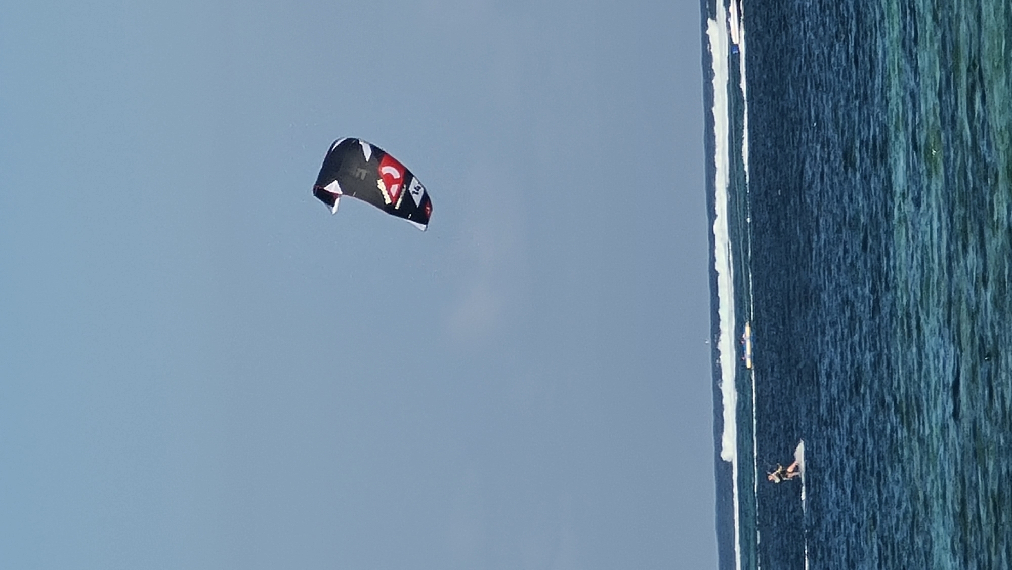 An individual kitesurfs on a body of water near the coast of Denpasar, Indonesia. The kitesurfer is positioned to the left of the frame, actively engaged in the sport. Above, a large, black and red kite is suspended in the clear blue sky, appearing to be controlled by the kitesurfer. The ocean is a deep blue, with white waves breaking in the distance, indicating a reef or shallow area. The water closer to the foreground is a lighter, translucent turquoise, suggesting a shallow reef. In the background, a small boat can be seen in the distance. The sky is largely clear with a few faint clouds.