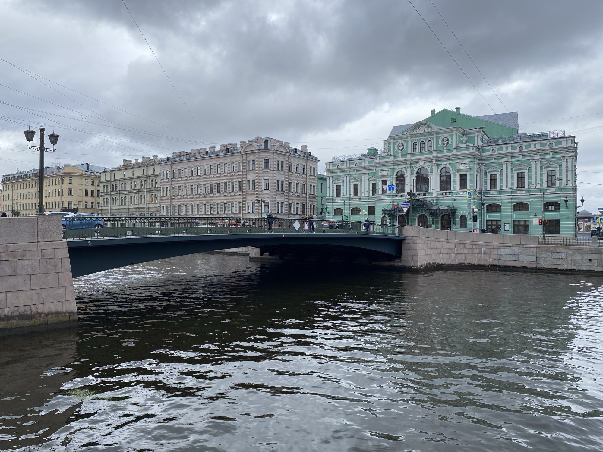 The image shows a view of the Griboyedov Canal in Saint Petersburg, Russia, on a cloudy day. A bridge spans the canal, with buildings lining the banks. On the right side of the canal, a light green building with ornate white trim, likely a theater or public building, is prominent. To its left is a lighter-colored, more utilitarian building. Several people are visible on the bridge and sidewalks. The water of the canal is dark and choppy, reflecting the overcast sky. Overhead, power lines and streetlights are visible.