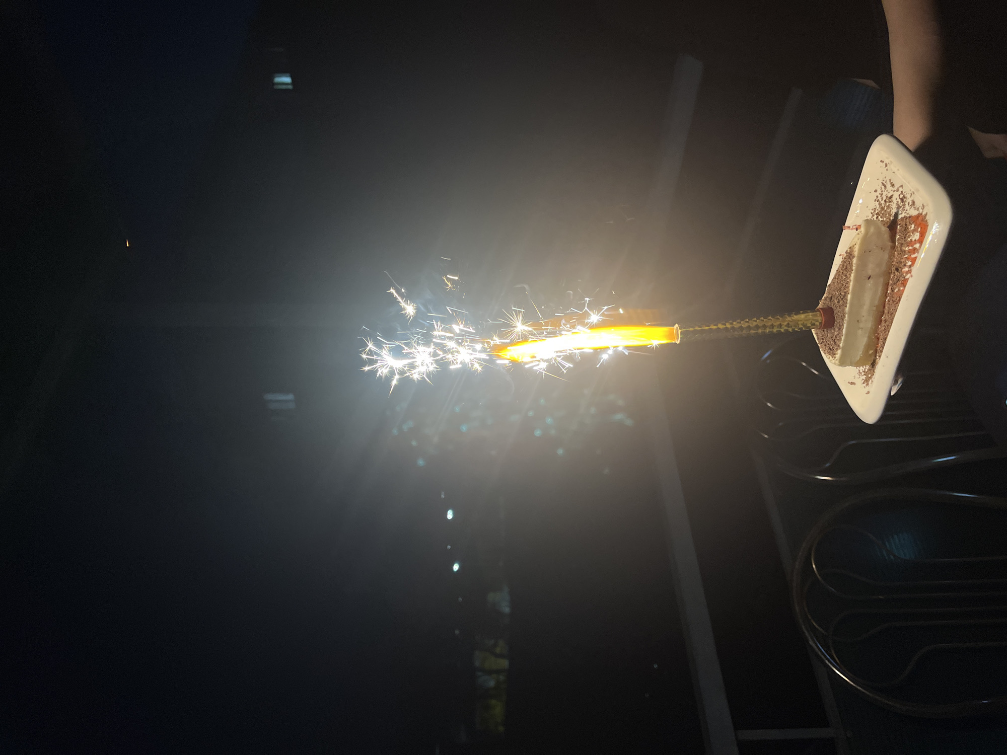 A sparkler emits sparks beside a slice of cake on a plate, held in someone's hand, possibly outdoors at night. The location is unknown; the city of Chaikovsky, Russia, cannot be confirmed based on the image. The plate is white with decorations, and the cake is light-colored. A dark background is visible behind the subject.