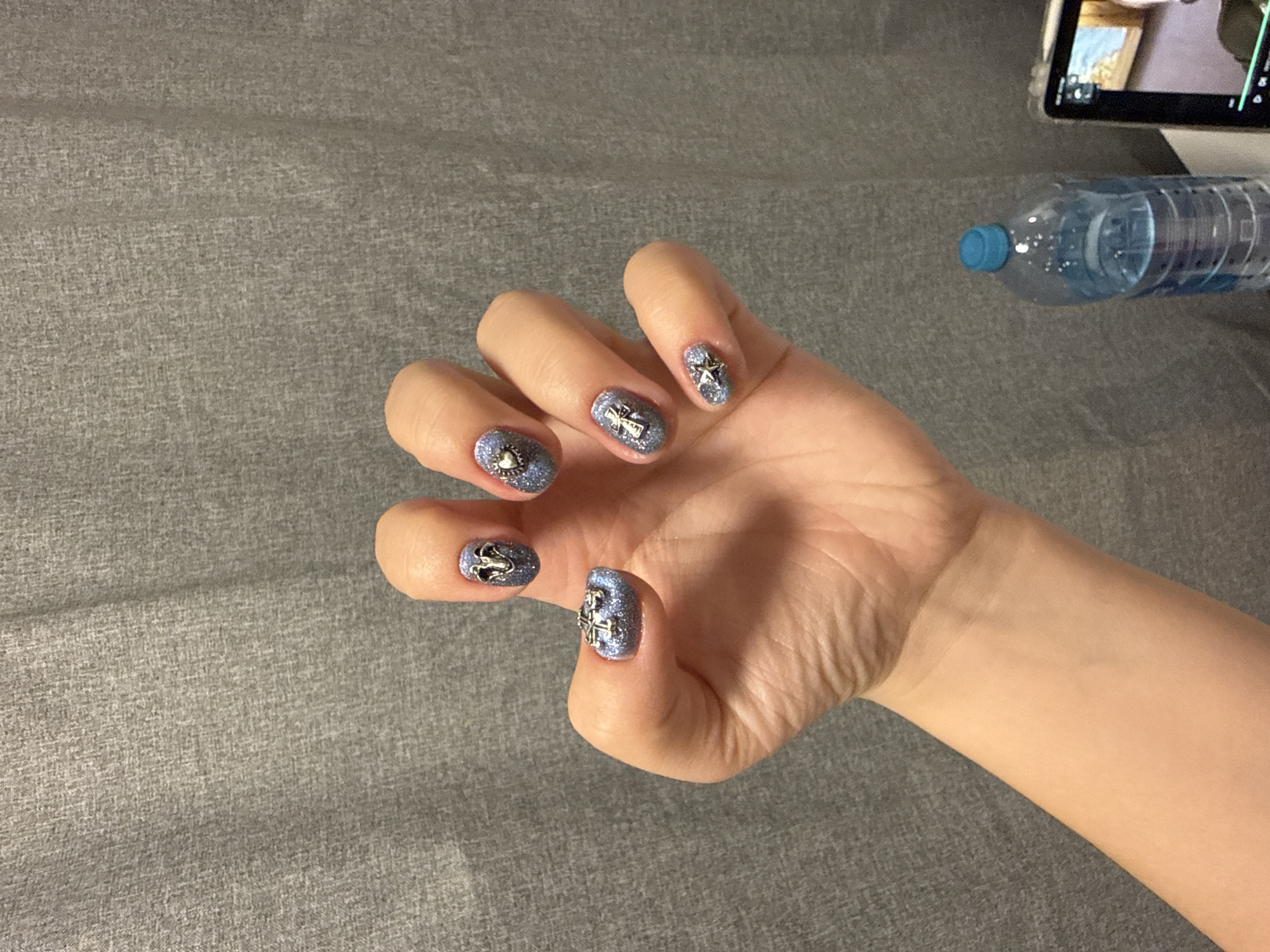 A human hand is positioned in the center-right of the frame, displaying five fingernails adorned with a light blue glitter polish and silver-colored embellishments. From left to right, the embellishments include a heart, a swan, a cross, a star, and a four-petal flower. The hand rests on a gray textured surface, possibly a fabric or upholstery. A partially visible clear plastic water bottle with a blue cap and a tablet or phone are situated in the upper right corner. The location is not identifiable as City of Zagreb, Croatia.