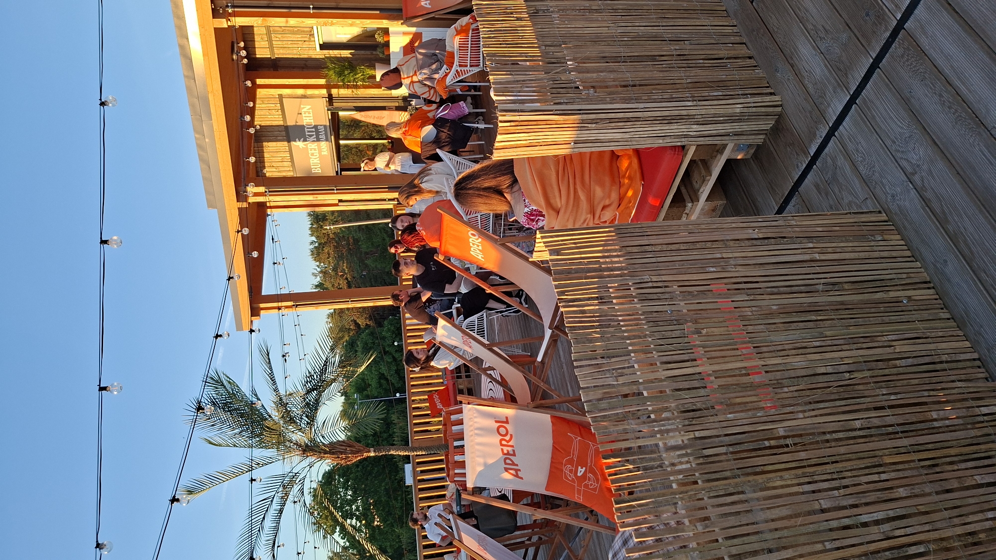 The image shows an outdoor seating area at a restaurant called "BURGER KITCHEN BANNABAR" in Tallinn, Estonia. It appears to be late afternoon or early evening, with the warm golden light of the setting sun casting long shadows and illuminating the wooden deck and bamboo screening. The sky is clear and blue.
Several people are seated in lounge chairs, some appearing to watch something, others conversing. Many are wrapped in blankets or wearing casual clothing, suggesting a relaxed atmosphere as the temperature cools. Some of the lounge chairs and flags display the "APEROL" logo, indicating it's likely a bar or restaurant that serves this aperitif. A palm tree is visible in the background, adding a touch of exoticism to the scene, which is set on a wooden deck, possibly near a beach or waterfront. The overall mood is one of leisure and enjoyment, with people unwinding and socializing outdoors.