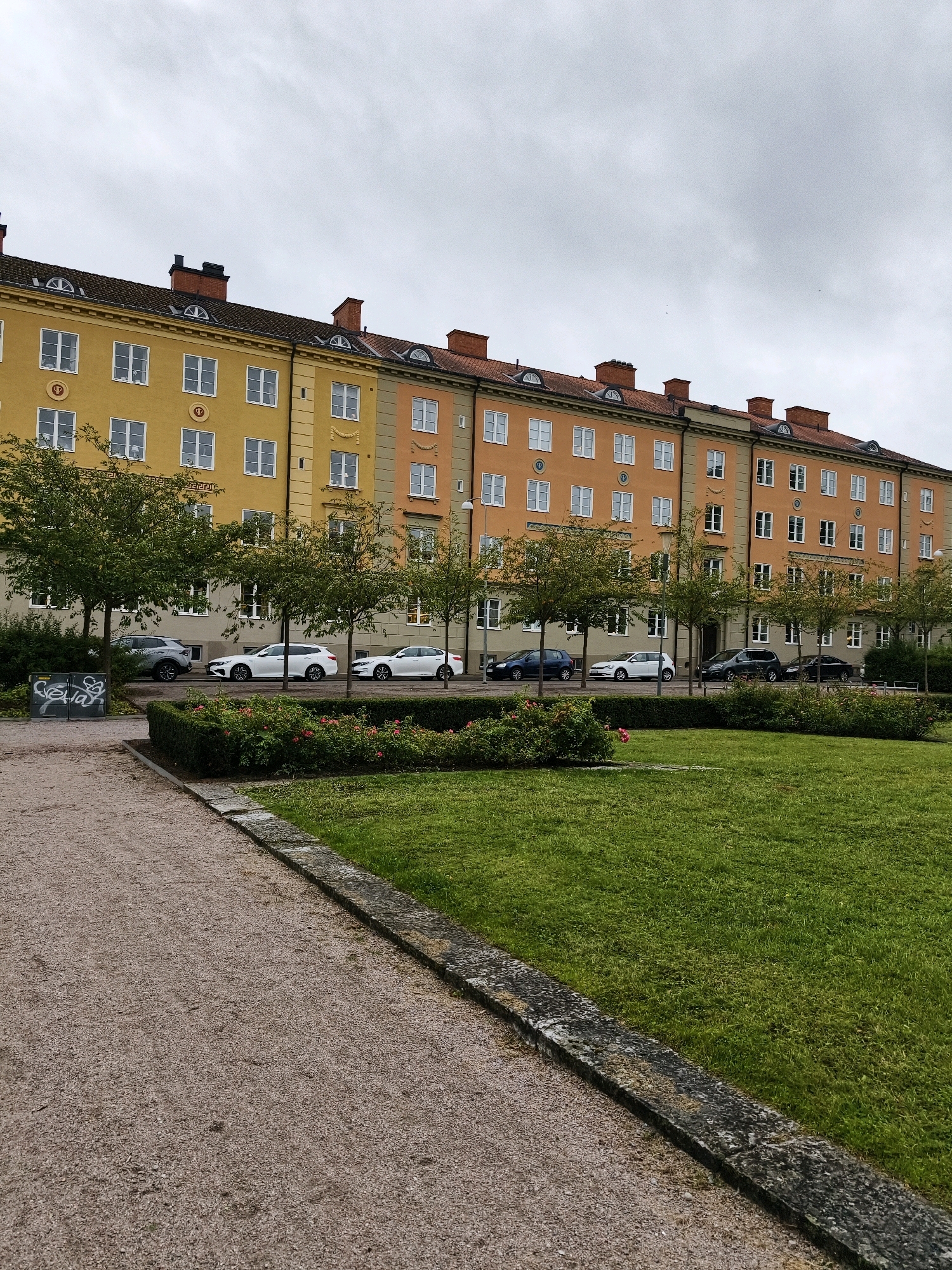 A row of colorful apartment buildings, painted in shades of yellow and orange, line a street in Linköping, Sweden. The buildings feature red-tiled roofs and numerous white-framed windows. Several cars are parked along the street in front of the buildings. A lush green lawn and a gravel path are visible in the foreground, separated by a stone border. The sky is overcast, suggesting a cloudy day.