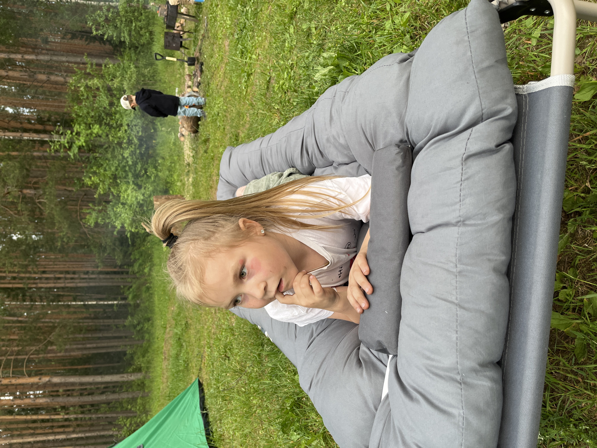 In Chaykovsky, Russia, a young girl with blonde hair pulled into a ponytail rests on a gray cot, nestled in a sleeping bag. She wears a white t-shirt and appears to be looking to her right, with her hand near her mouth. Her expression is thoughtful or perhaps slightly sleepy.
In the background, a small campfire smolders with smoke rising, suggesting a camping or outdoor gathering. A person in a dark hoodie and light-colored pants stands near the fire pit, facing away from the camera. There are also some logs and camping equipment visible near the fire, including what looks like a shovel. To the left of the frame, a green tent is partially visible. The setting is outdoors, with a lush green grassy area and a dense forest of pine trees in the background. The lighting suggests it is daytime, possibly late afternoon or early evening. The overall atmosphere is relaxed and suggests a family or group enjoying time in nature. No visible text is present in the image.