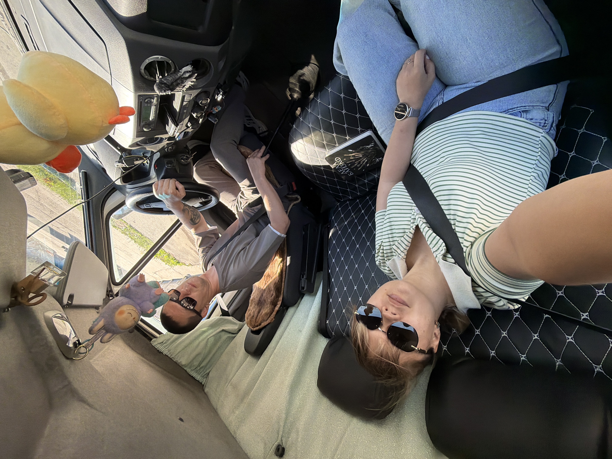 In a camper van in Chaikovsky, Russia, a man in a grey t-shirt and sunglasses is driving. A woman in a green and white striped shirt and blue jeans is lying down in the passenger seat, also wearing sunglasses. They are both wearing seatbelts. A plush chicken toy hangs from the ceiling. A book with Russian text is on the seat between them. The van appears to be parked on a street with some greenery visible outside.