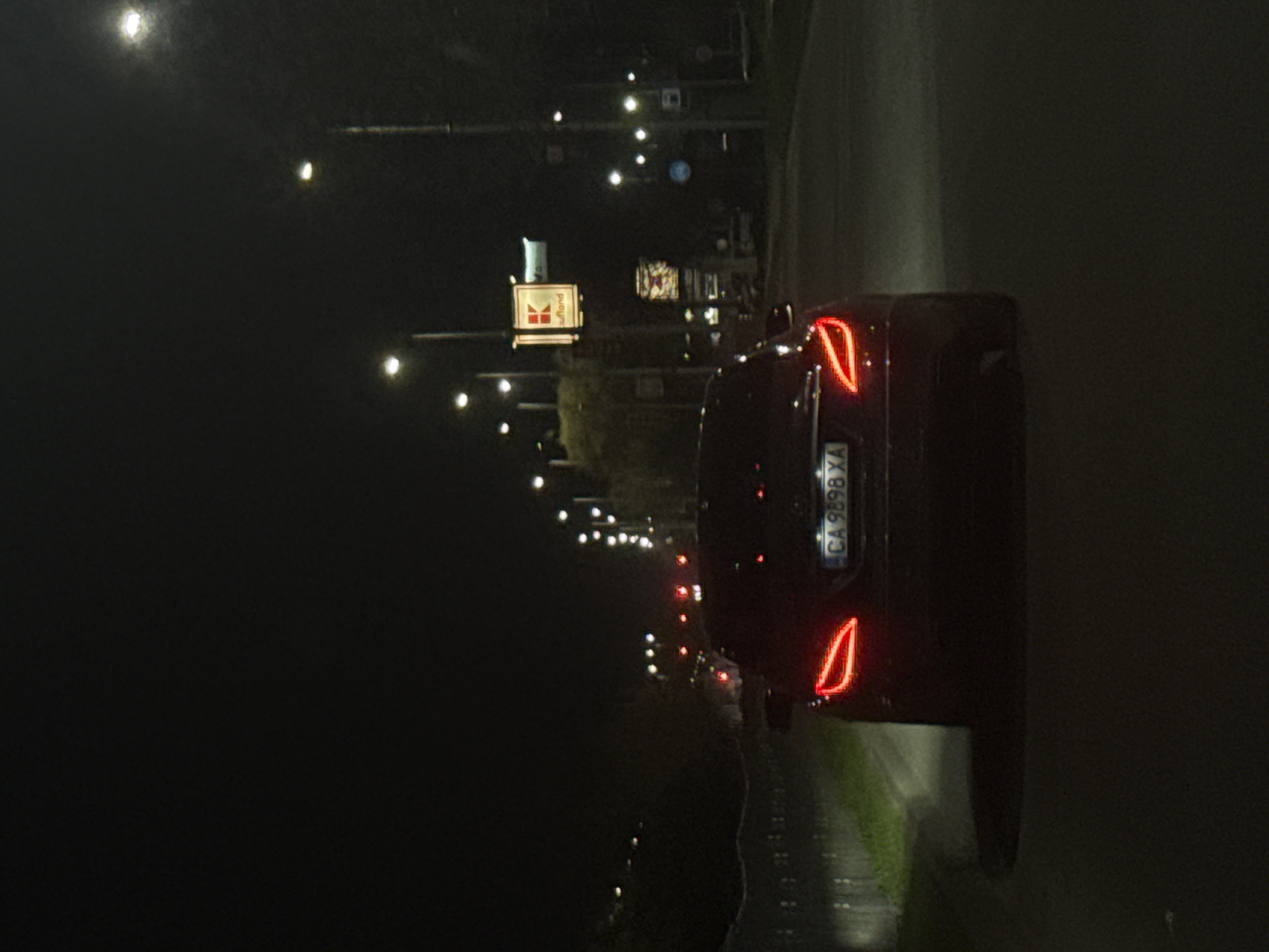 The image shows a night scene in Plovdiv, Bulgaria, featuring a dark-colored car on a road. The car's license plate reads "CA 9898 XA." Streetlights illuminate the area, and a sign with the Kaufland logo is visible in the background. The environment appears to be a road with surrounding vegetation.