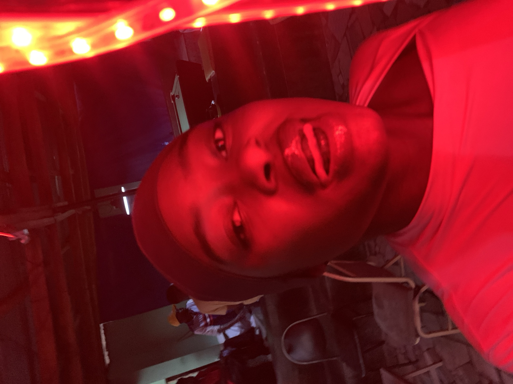 The image shows a person illuminated by a red light source, possibly a string of lights, casting a strong red tint over the entire scene. The person has dark skin and is wearing a dark-colored head covering. Their lips are slightly parted. In the background, there are other people, one wearing a yellow hat and light-colored clothing, seemingly seated. Objects in the background include chairs and structural elements, possibly walls or curtains. The location is said to be in New Stadium, Nigeria, though this cannot be confirmed visually from the image alone. The setting appears to be indoors, possibly a club or bar. The overall mood is dark and saturated with red light.