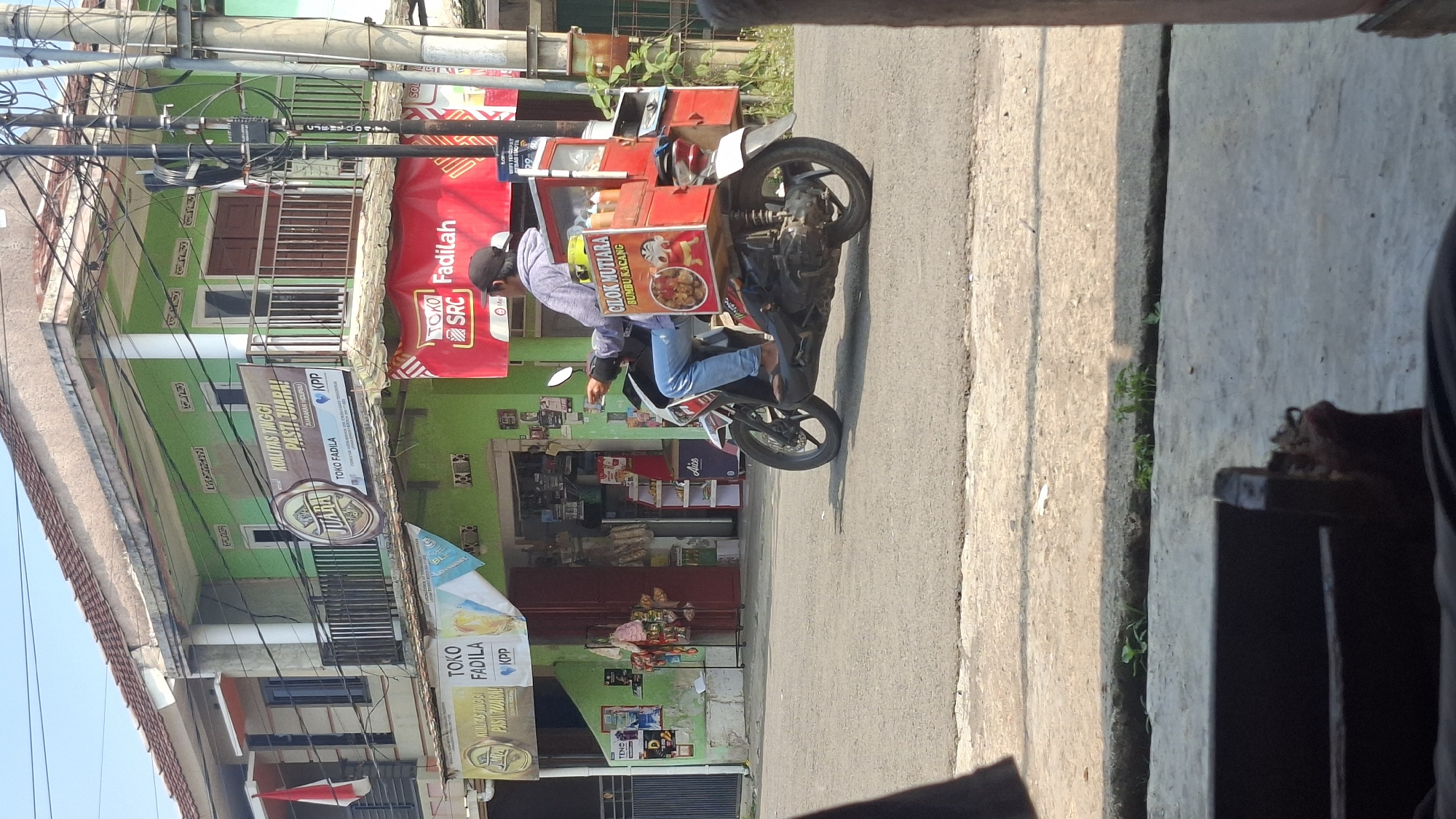 A man in a gray hoodie and blue jeans rides a scooter on a paved road in Cilaku, Indonesia. The scooter has a red food cart attached to the back, displaying a picture of a cartoon character and the text "CILOK MUTIARA BUMBU KACANG." The man is wearing a black baseball cap and is looking forward. Behind him, there is a two-story building painted green, with a balcony on the second floor. Several signs are visible on the building, including one that reads "KUALITAS TINGGI PASTI JUARA" and "TOKO FADILA." Another red banner with "TOKO SRC Fadilah" is also visible. The environment appears to be a street scene with buildings and utility poles. The lighting suggests it is daytime, possibly morning or afternoon.