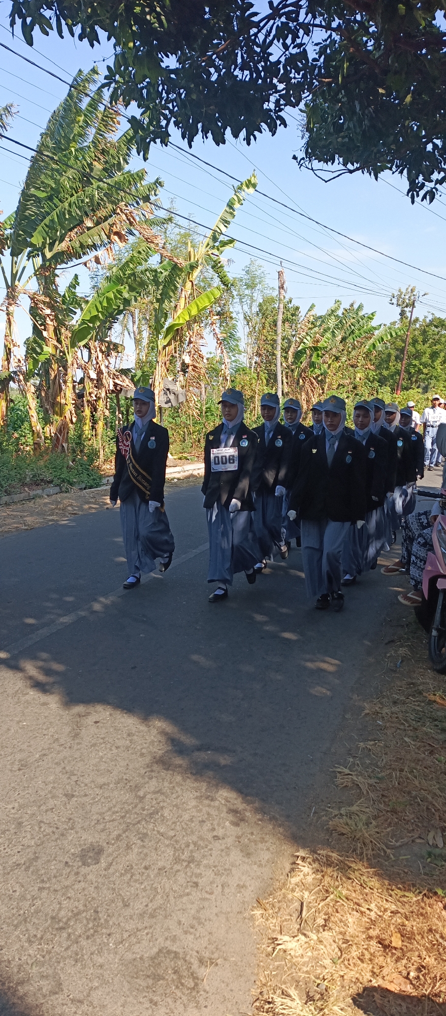 In Ketowan, Indonesia, a group of young people, likely students, are marching in a procession on a paved road. They are dressed in dark blazers, light gray pants, white shirts, and caps. Some wear sashes across their chests. One individual in the front left wears a dark sash with white lettering that is partially obscured. Another individual in the front center wears a bib with the number "008" printed on it. They are walking in formation, appearing to be participating in a parade or ceremony. The environment is outdoors, with a clear blue sky overhead and lush green foliage, including banana trees, lining the side of the road. The road is asphalt with a white dividing line. The lighting suggests it is daytime.