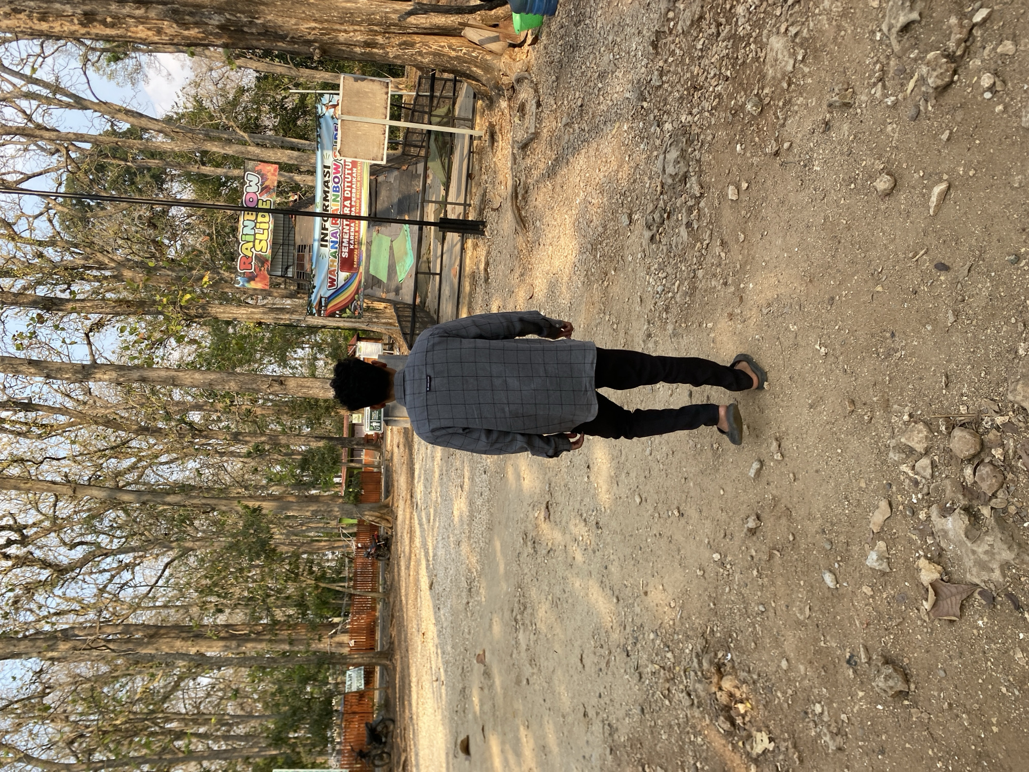 A person walks away from the camera on a dusty path in a park with tall trees. The person is wearing a grey plaid long-sleeved shirt and black pants. In the background, there is a rainbow slide attraction with a banner that reads "INFORMASI WAHANA RAINBOW SLIDE". There are also a few motorcycles parked near a wooden fence. The ground is covered in dirt and rocks, and the trees are bare, suggesting it may be a dry season. The location is identified as Ketileng, Indonesia.