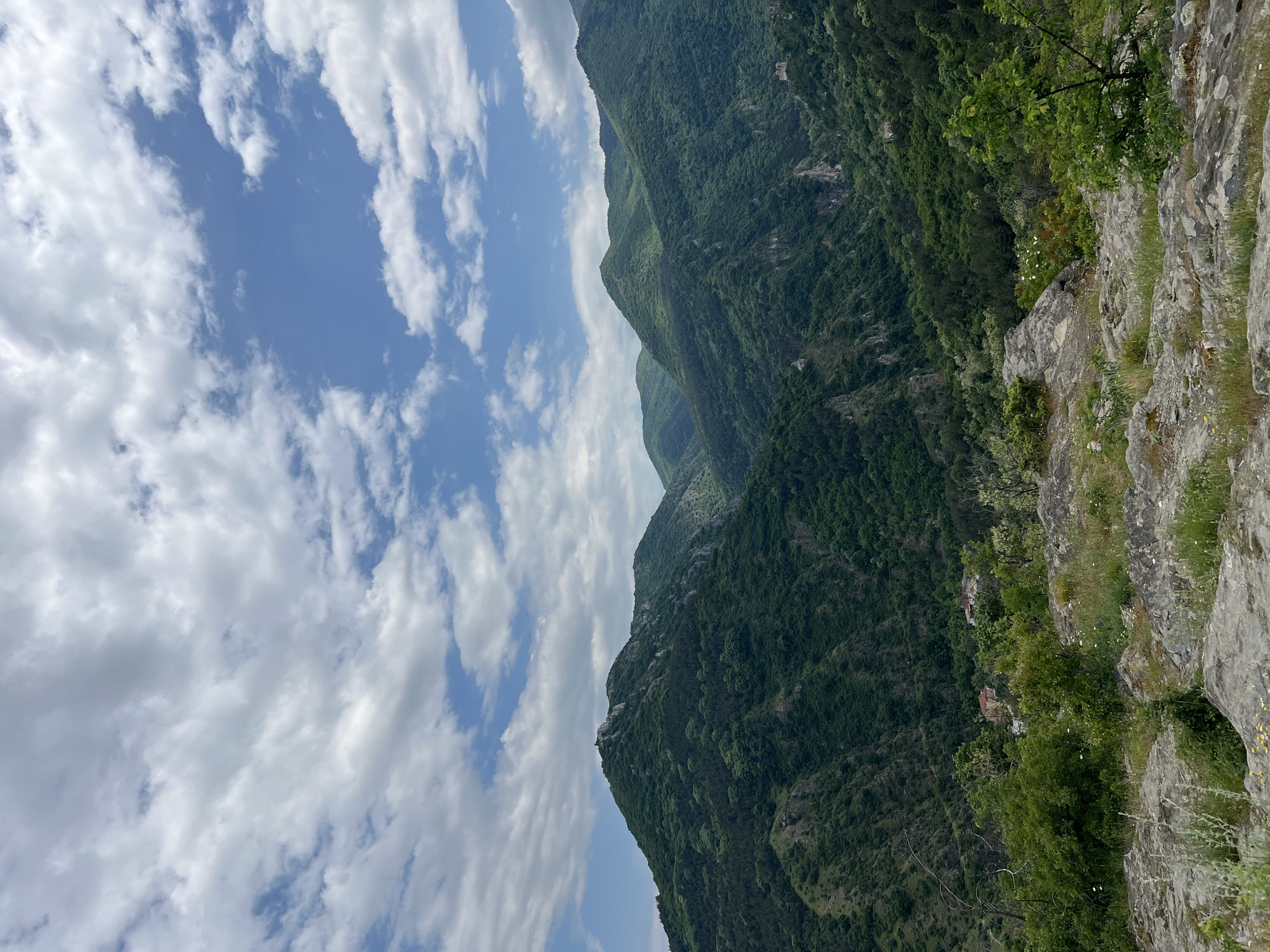 The image shows a view of a mountainous landscape. The sky is blue with scattered white clouds. In the foreground, there are rocky outcrops and greenery. The mountains are covered in lush green vegetation, suggesting a forested area. A few buildings are visible nestled within the foliage on the mountainside. The image was taken from a high vantage point overlooking the terrain, likely near the city of Asenovgrad, Bulgaria, based on the typical landscapes of the area.