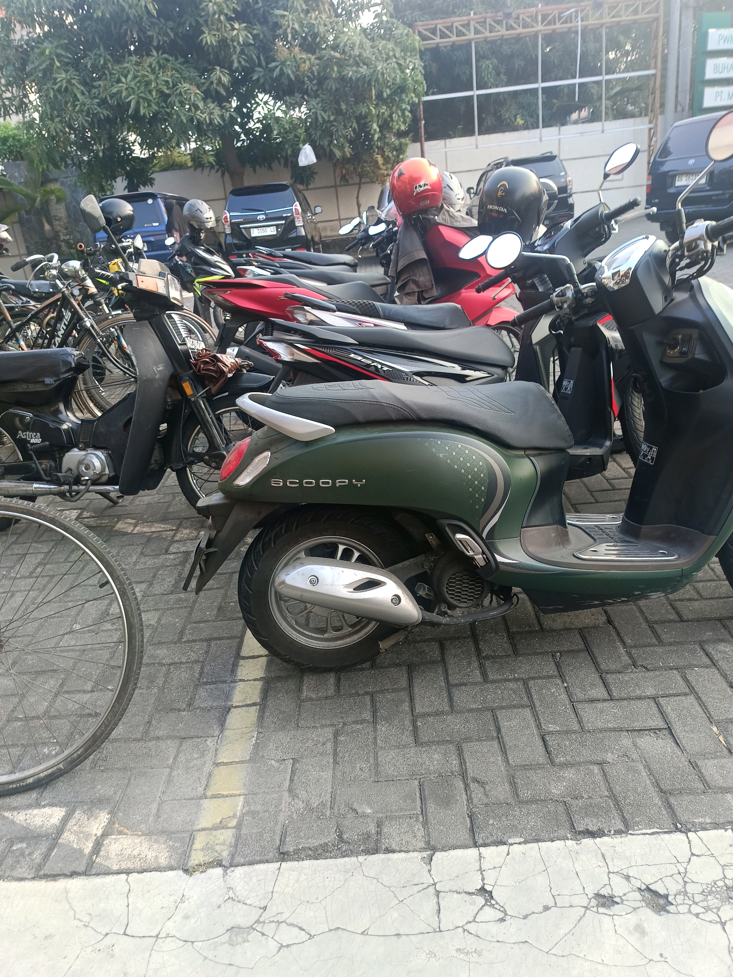 This is a photograph of a street scene in Yogyakarta, Indonesia, likely on a sunny day. The foreground is dominated by parked motorcycles and a bicycle. A green Honda Scoopy scooter is prominently featured on the right, its matte green body contrasting with the paved brick surface. To its left, a black motorcycle with red accents is parked, and further left, a bicycle with a visible silver rim and spokes occupies the bottom left corner.
In the background, several other motorcycles are parked, some with helmets on their seats. Behind the motorcycles, a dark blue car is visible, followed by a lighter blue vehicle. Trees with dense green foliage line the area behind the vehicles, and a building structure with a metal roof and railing is present in the upper background. A portion of a sign board is visible on the far right, with some text that is not entirely legible. The ground is paved with interlocking bricks, with a white painted line running across it. The overall impression is that of a parking area for two-wheeled vehicles, common in urban Indonesian settings.