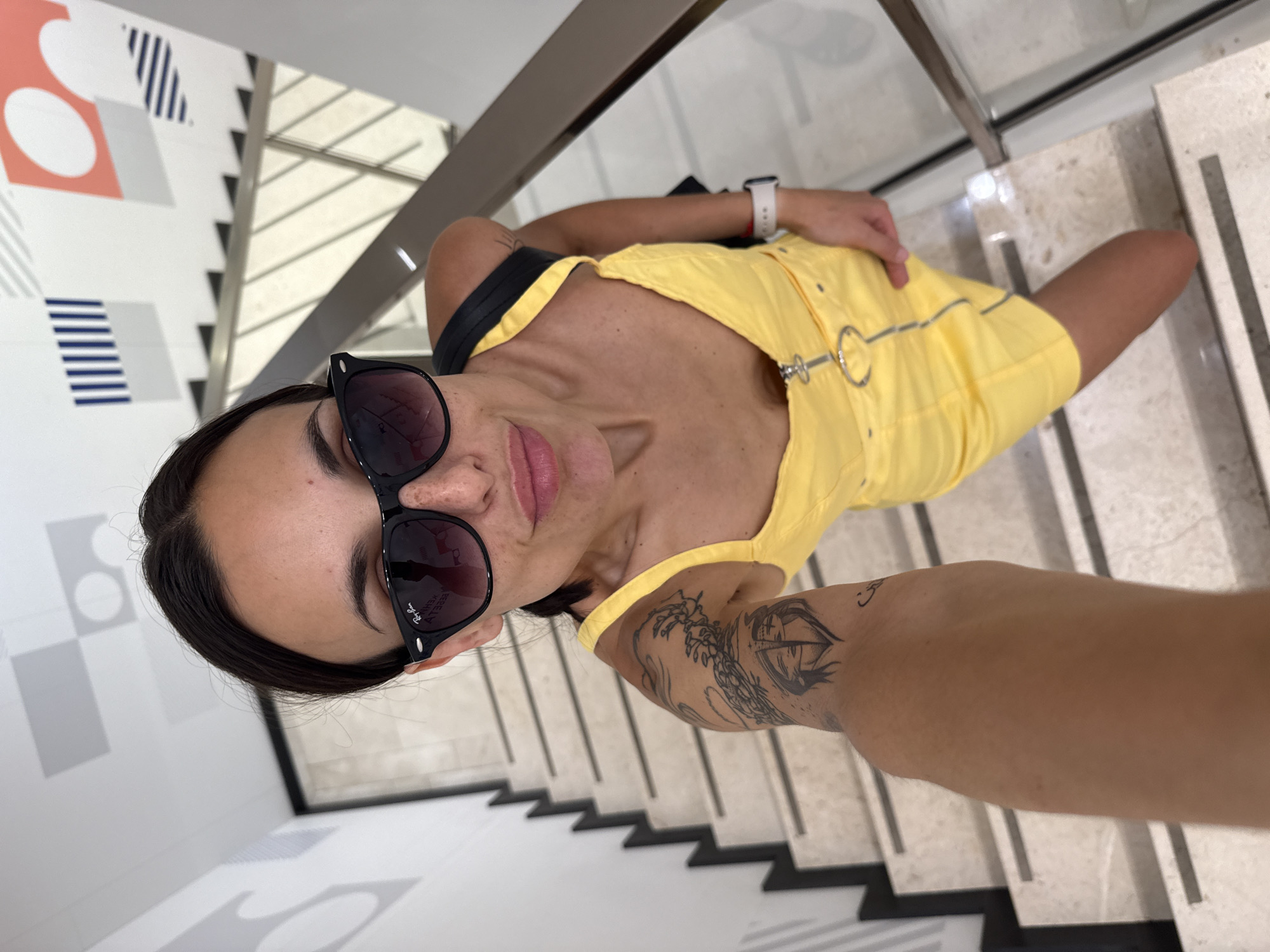 A person, appearing female, is the central subject of a selfie taken from an elevated angle, looking up at the camera. They are dressed in a bright yellow mini-dress or romper, featuring silver ring and stud details on the front. Dark sunglasses obscure their eyes, with the "Ray-Ban" brand name visible on the temple, along with mirrored, partially legible text reflecting on the lenses. A white-banded smartwatch is worn on the right wrist, and a black strap, possibly from a bag, crosses their left shoulder. Their left arm, extended to hold the camera, displays several tattoos, including a prominent large eye design and a smaller symbol near the elbow.
The setting appears to be a modern indoor environment, likely in Pleven, Bulgaria. The person is positioned on a sleek staircase, characterized by light-colored treads and dark accents. In the background, a wall features abstract geometric patterns composed of orange, white, grey, and blue shapes and stripes, contributing to a contemporary aesthetic. Metallic or reflective railings are also visible, hinting at a public or commercial building. The lighting is bright and even, suggesting an indoor daytime setting. The person maintains a neutral to slightly smiling expression.